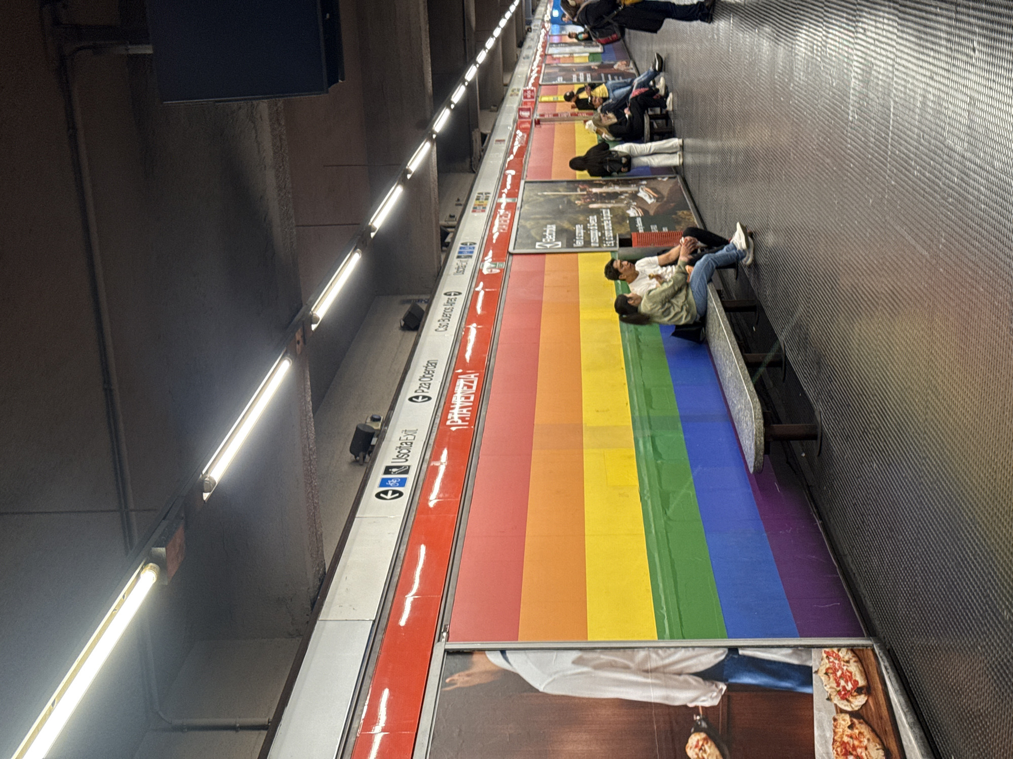 This image depicts a subway or metro station platform, likely in Milan, Italy, given the visible Italian text. The long platform is illuminated by numerous rectangular fluorescent lights running along the ceiling. The floor features a distinctive dark gray, textured surface.
A significant portion of the platform wall is covered by a vibrant, multi-colored banner displaying the full spectrum of the rainbow flag (red, orange, yellow, green, blue, and purple). This prominent banner stretches horizontally, serving as a bright backdrop to the scene. Interspersed with and below the rainbow banner are various advertisements. One ad clearly shows the brand "Electrolux" and includes Italian text. Closer to the foreground, another advertisement features a person and what appear to be pizzas.
Several people are visible on the platform, mostly seated on metal benches positioned in front of the colorful wall and advertisements. They appear to be commuters or travelers waiting for a train. Some individuals are looking down, possibly at their phones, while others are resting. One person in the foreground wears a light green jacket and blue jeans, sitting with their feet up on the bench. Further down the platform, more people are seen in various postures of waiting.
Above the rainbow banner, a white strip contains directional and informational text written in Italian. Visible phrases include "Uscita Exit" (Exit), "P.zza Oberdan", "1^ PRIMAVERA", "P.zza Doman", and "Corti Baires". The text "1^ PRIMAVERA" is notably larger and printed in red. The artificial lighting suggests an indoor setting where the time of day is not discernible.