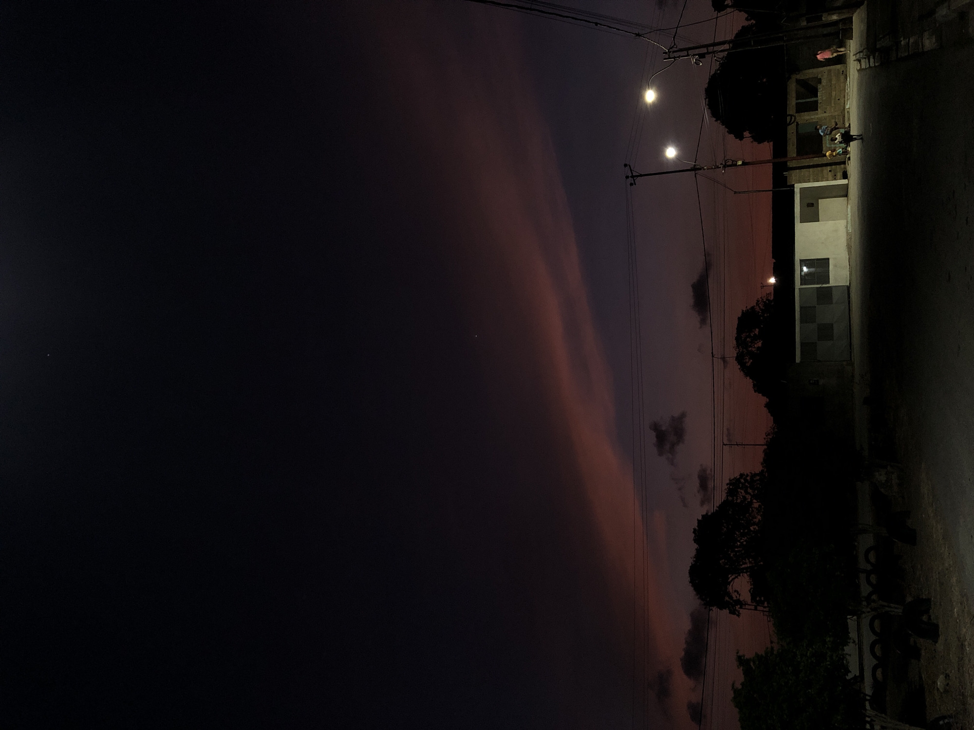 A dimly lit, horizontally-oriented photograph captures a street scene in Altagracia, Venezuela, at dusk or night. The sky transitions from a deep black on the left to a mix of purple and orange hues, indicating the sunset or sunrise. Power lines run across the upper portion of the sky, intersecting with two illuminated streetlights. On the right side of the frame, a single-story building stands adjacent to the street. Individuals appear to be gathered outside the building. The foreground shows a road surface. Silhouetted trees are scattered throughout the middle ground of the image, and tire piles lie closer to the roadside.