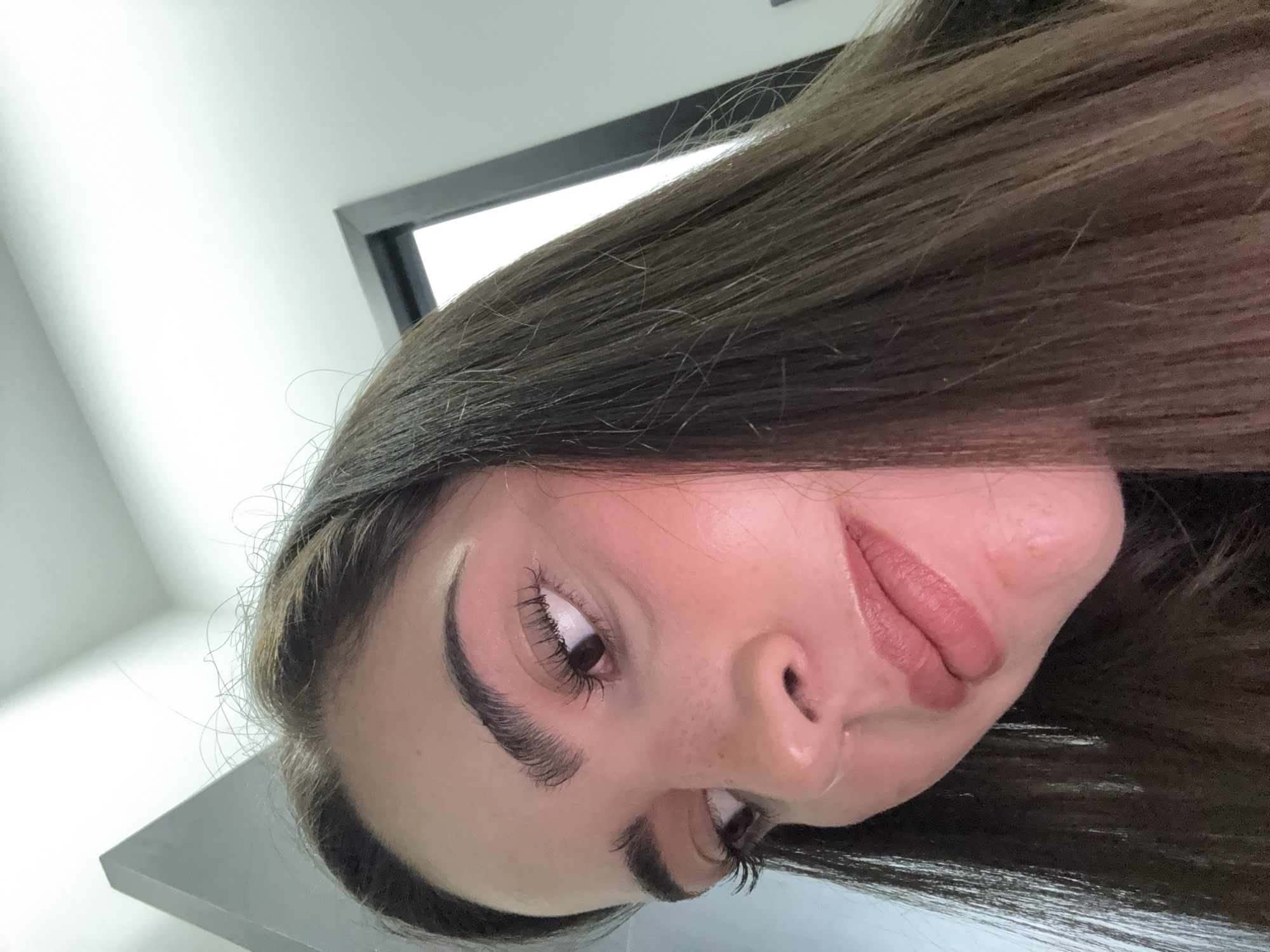 The image shows a close-up view of a person's face and hair, tilted to the side. The person has brown hair, fair skin, and prominent eyebrows. Freckles are visible on the nose. The person is wearing lip makeup. The background includes a white wall and a black-framed opening. The location is not discernible from the image. The image does not show any elements that can be used to identify the city of San Bernardino Tlaxcalancingo, Mexico.