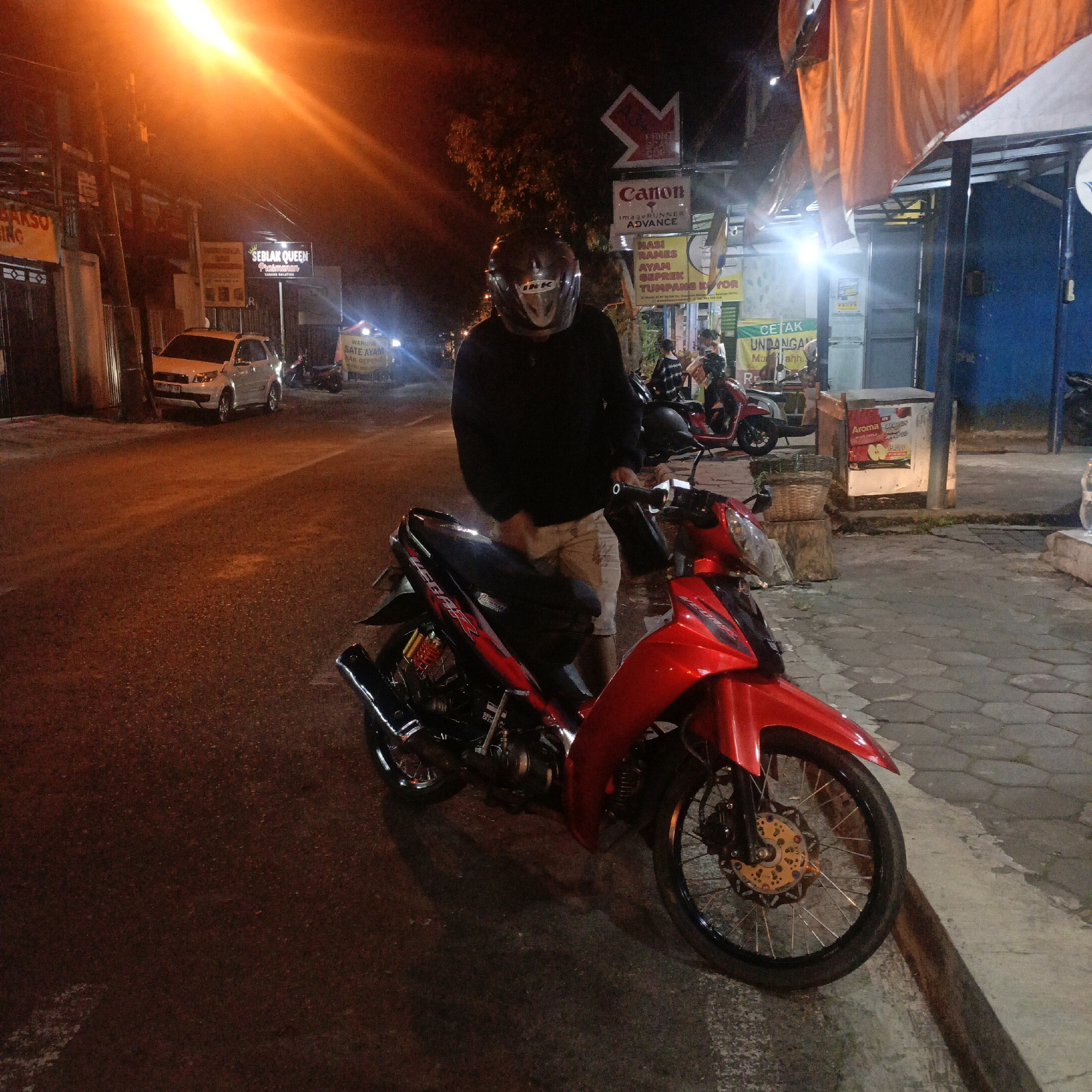 The image depicts a nighttime street scene in Salatiga, Indonesia. In the foreground, a person wearing a dark long-sleeved shirt, light-colored shorts, and a helmet stands next to a red motorcycle. The motorcycle is the primary subject, featuring a black seat, red body panels, and gold-accented wheels. The person appears to be interacting with the motorcycle, possibly preparing to ride it.
The background reveals elements of an urban environment. To the left, a white car is parked on the side of the road, and beyond it, a building with illuminated signage is visible, including one that reads "SEBLAK QUEEN PRASMANAN" and another displaying "BAKSO KING". A street lamp casts a bright orange glow over the scene, illuminating the street and the surrounding structures. Further back, other motorcycles and individuals are present, along with various shopfronts and signs, suggesting a commercial or entertainment area. The ground is a mix of asphalt road and a paved sidewalk. The overall lighting and atmosphere indicate it is nighttime, with artificial lights dominating the scene.