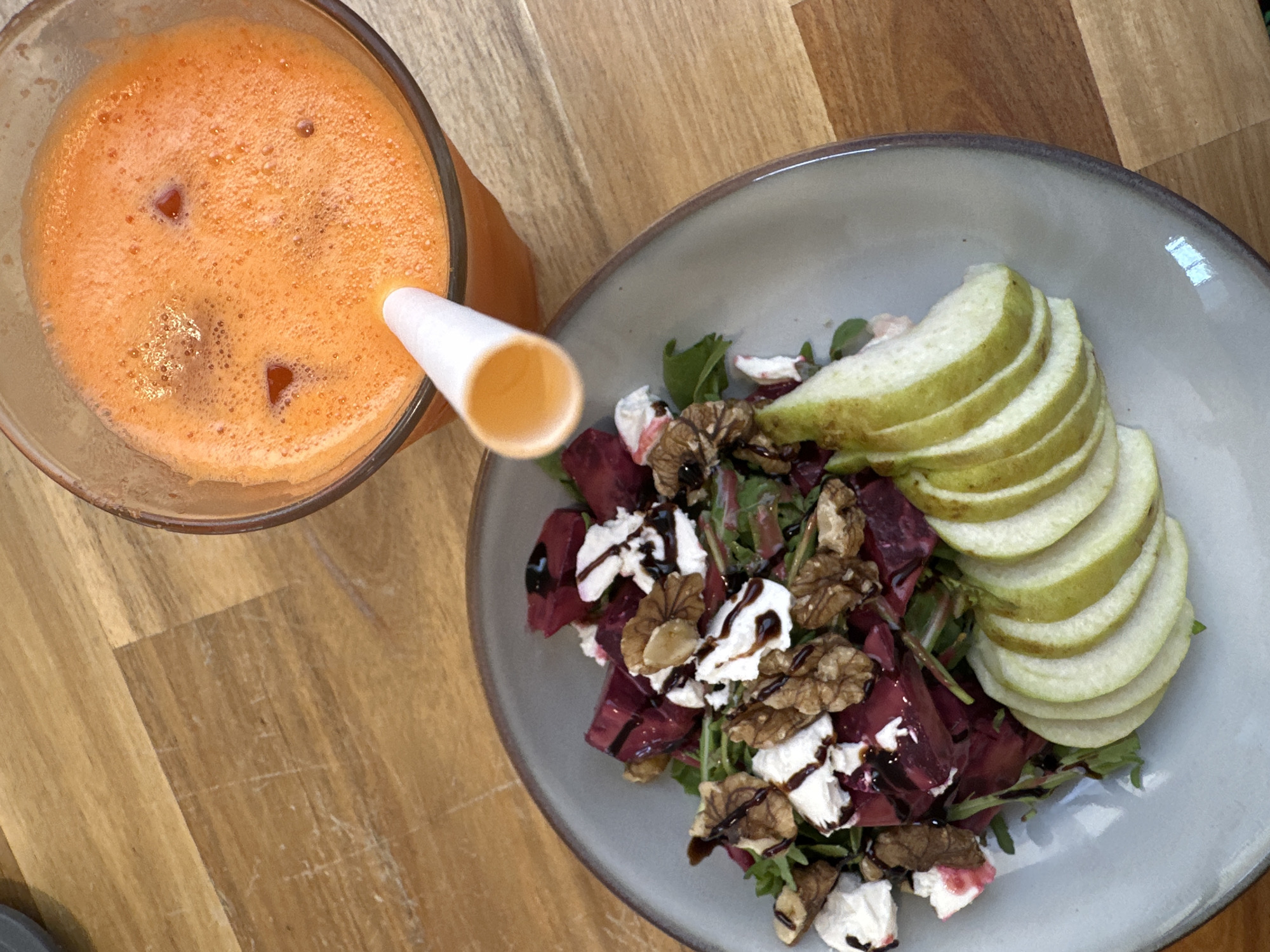 The image shows a close-up, overhead view of a meal and a drink on a wooden table. To the right, a glass filled with vibrant orange juice, likely carrot juice, sits with ice cubes and a white straw. To the left, a plate contains a salad composed of deep red beets, crumbled white cheese (possibly goat cheese or feta), walnuts, and dark green leafy vegetables. Sliced pear is arranged on the side of the plate. A dark drizzle, possibly balsamic glaze, is visible over the salad. The setting appears to be a restaurant or cafe in ตำบลเกาะพะงัน, Thailand.