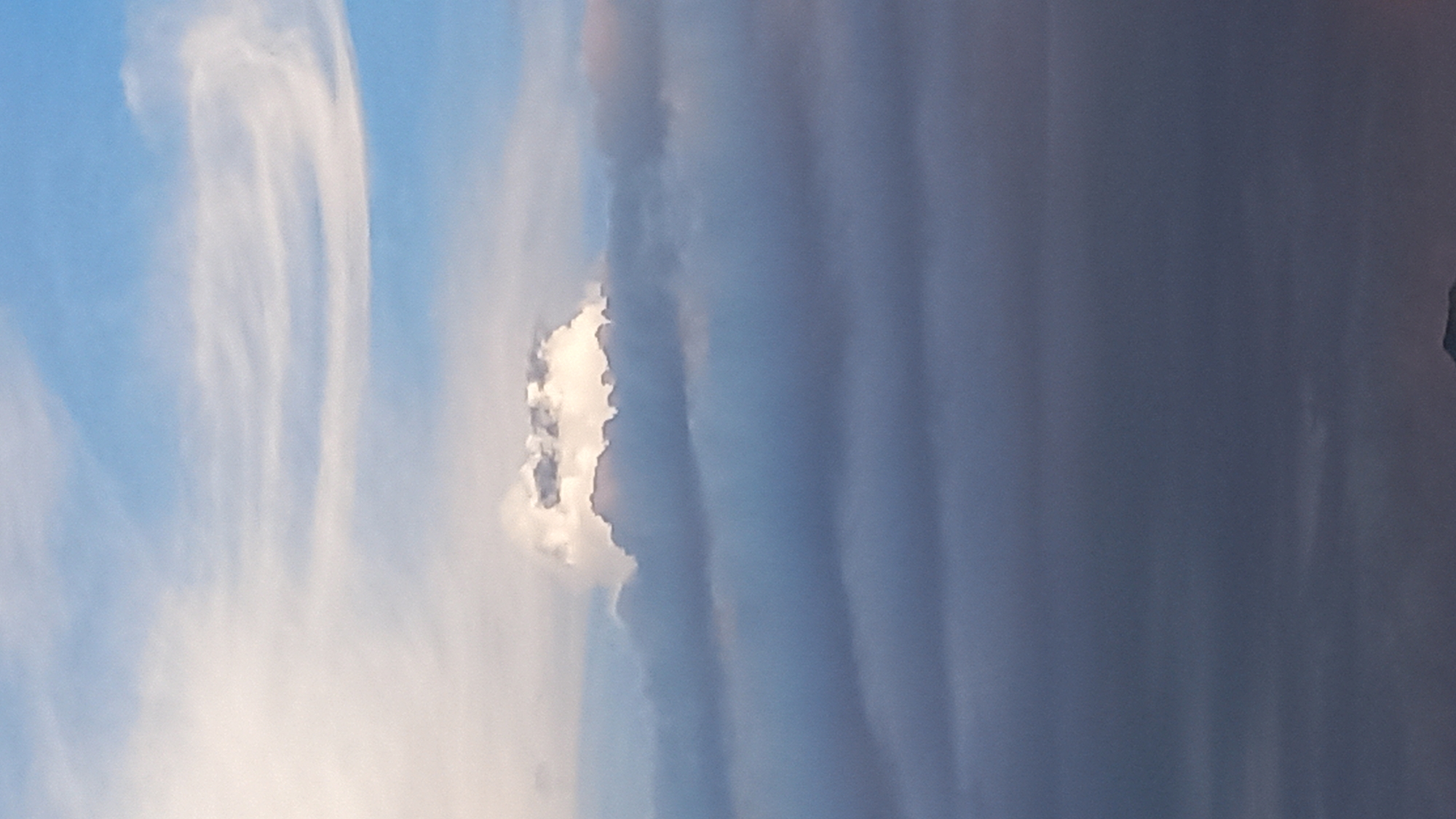 The image shows a layered cloud formation against a light blue sky. The foreground is dominated by dark, dense clouds, with a few lighter, wispy clouds appearing above them. In the upper portion of the image, a bright, illuminated cloud stands out against the darker clouds beneath it, suggesting sunlight piercing through. The top of the sky shows thinner, streaky clouds against a clear blue backdrop. The bottom edge of the image contains a hint of what appears to be a dark, silhouetted landmass, possibly mountains or a hilly terrain. The overall scene depicts a dynamic sky with varying cloud densities and lighting conditions, typical of weather formations above a location like Koh Phangan, Thailand, where cloudscapes can be dramatic and varied.