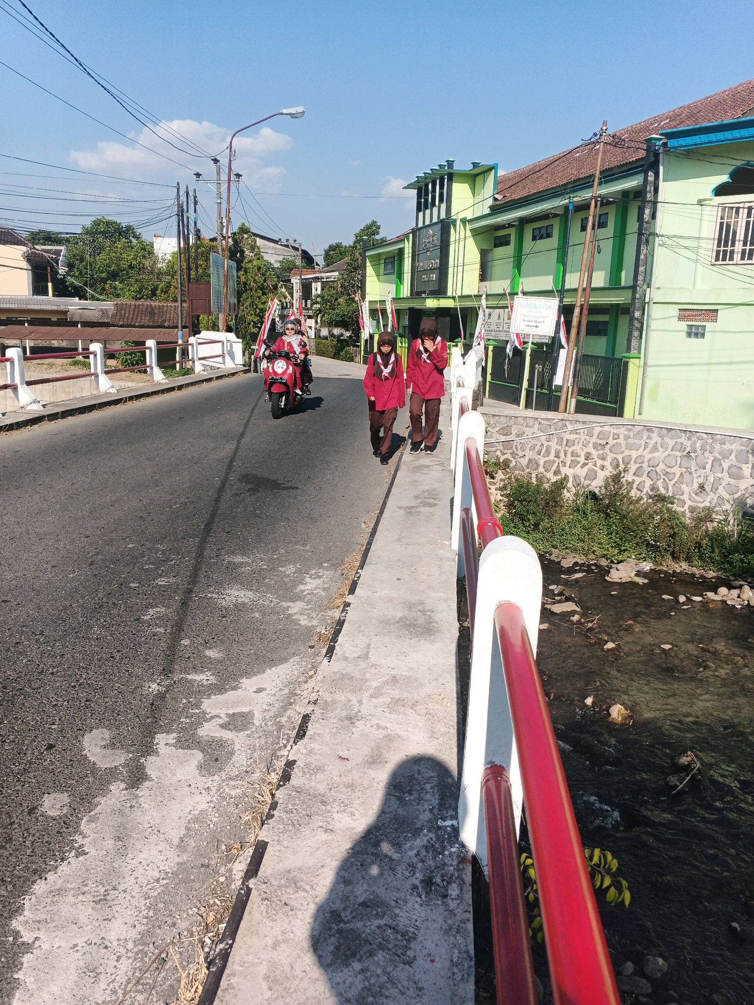 The image shows a street scene in Ungaran, Indonesia, on a sunny day. In the foreground, a concrete sidewalk runs alongside a road. A red and white railing borders the sidewalk, separating it from a shallow, rocky stream below. The road is paved and has some water stains. Two young people wearing maroon uniforms and headscarves are walking on the sidewalk, facing away from the camera. A red motorcycle with two occupants, also wearing similar uniforms, is driving on the road. In the background, a light green building with a red-tiled roof and white window frames is visible. The building has some signage and flags in front of it. Trees and utility poles line the street, and the sky is clear blue with a few scattered clouds. The scene appears to be a quiet street in a residential or school area.