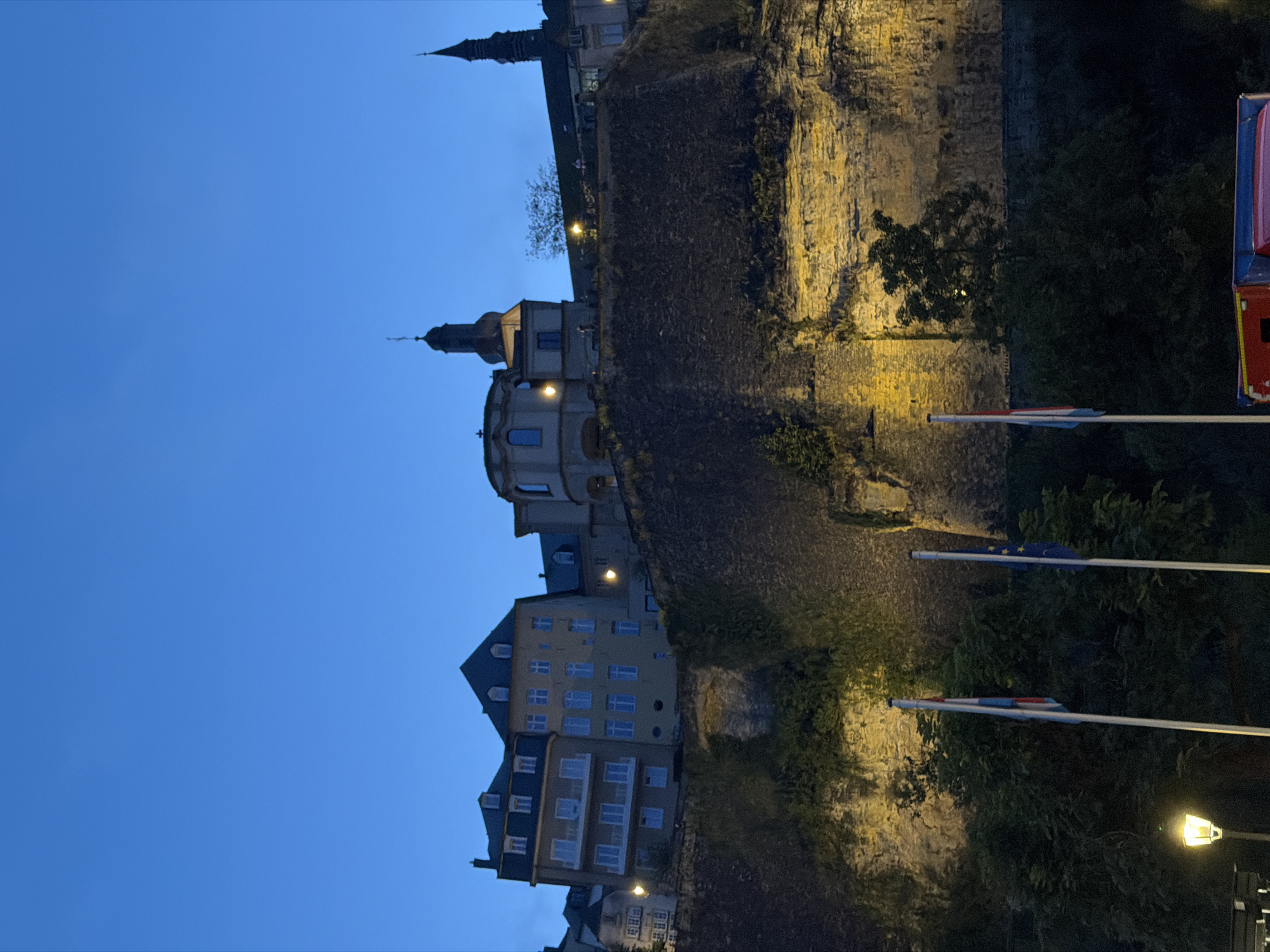 This photograph captures a twilight view of the Bock Casemates in Luxembourg City, Luxembourg. The foreground features dark green foliage and a lamp post with a warm yellow light. Three flags are prominently displayed on flagpoles: one with red, white, and blue stripes, another with blue and yellow stars, and a third flag which is partially obscured. Behind the flags, a large, imposing stone wall, characteristic of Luxembourg's historic fortifications, rises steeply. This wall is illuminated from below, casting a yellow glow on its textured surface and the vegetation clinging to it.
Perched atop the wall are several buildings, appearing in silhouette against the deep blue twilight sky. These include multi-story structures with windows and balconies, and taller buildings with spired rooftops, hinting at churches or other historical landmarks. The architecture suggests a blend of residential and possibly administrative or religious buildings. The overall scene evokes a sense of history and timelessness, with the ancient fortifications juxtaposed against the more modern buildings and the clear evening sky.