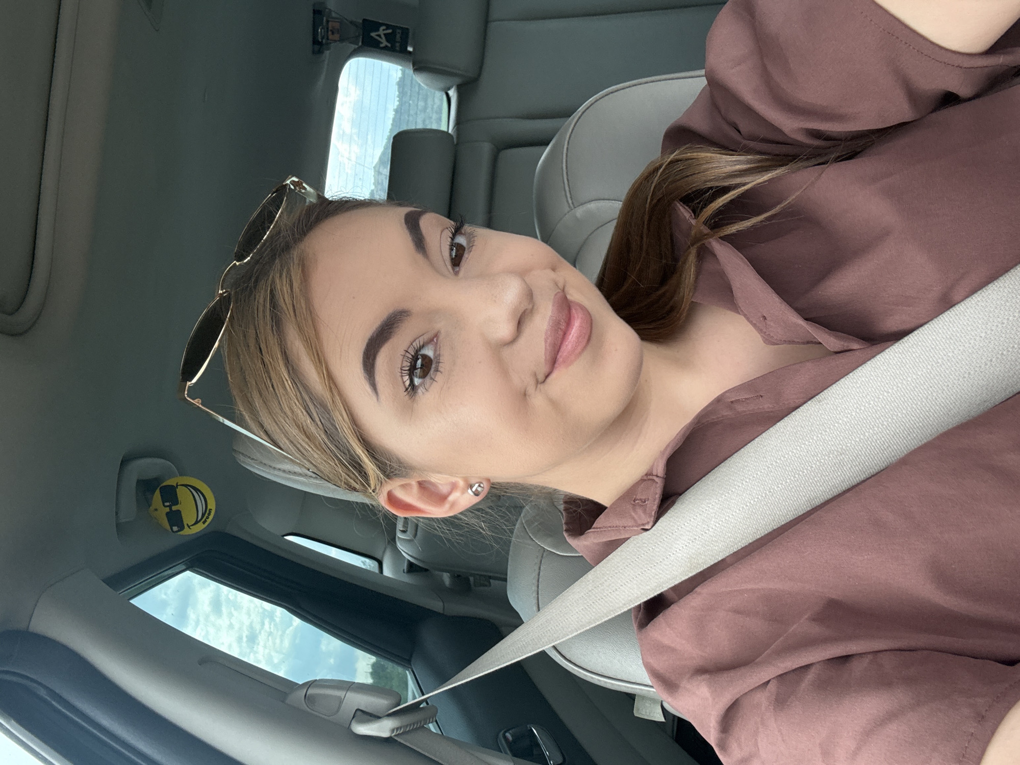 A young woman with brown hair pulled back and sunglasses perched on top of her head is taking a selfie inside a car. She is wearing a maroon collared shirt and a gray seatbelt. The interior of the car is visible, including the seats and the sky through the windows. A smiley face air freshener with sunglasses is hanging from the rearview mirror. The scene appears to be in Aldomirovtsi, Bulgaria.