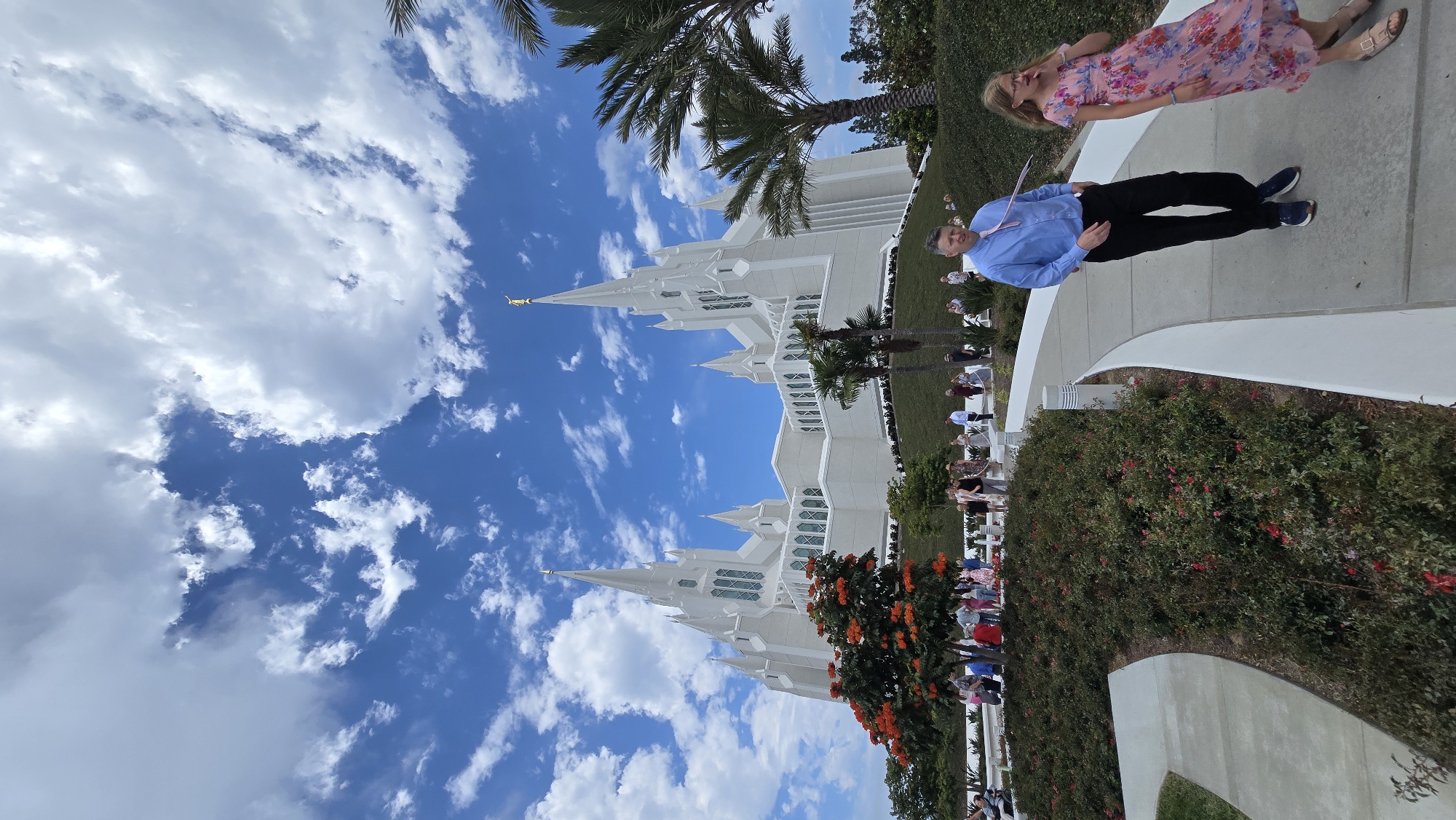 This media file captures a bright, sunny day with fluffy white clouds in a vibrant blue sky. The scene appears to be outdoors, possibly in a park or near a significant building. In the foreground, a group of people are visible. One man in a blue dress shirt and black pants stands near the center, looking towards the camera with a slight smile. To his left, a woman in a floral pink dress is also looking slightly away from the camera, with a relaxed expression. Other individuals are scattered throughout the frame, some sitting on benches and others standing.
Dominating the background is a large, white, ornate building with multiple spires, characteristic of a temple or similar religious structure. The architecture is detailed and grand, with pointed roofs and clean lines. Lush green landscaping, including trees and bushes with red flowers, surrounds the building and the walkway where the people are gathered. Palm trees are also visible, suggesting a warm climate.
The lighting indicates it's daytime, likely late morning or afternoon, given the brightness and the shadows. The overall mood seems pleasant and serene, with people dressed in casual to semi-formal attire. The image's orientation is sideways, with the sky appearing on the left and the ground on the right. There is no visible text in the image. The presence of the distinctive architecture, palm trees, and clear skies suggests a location in San Diego, California, United States.