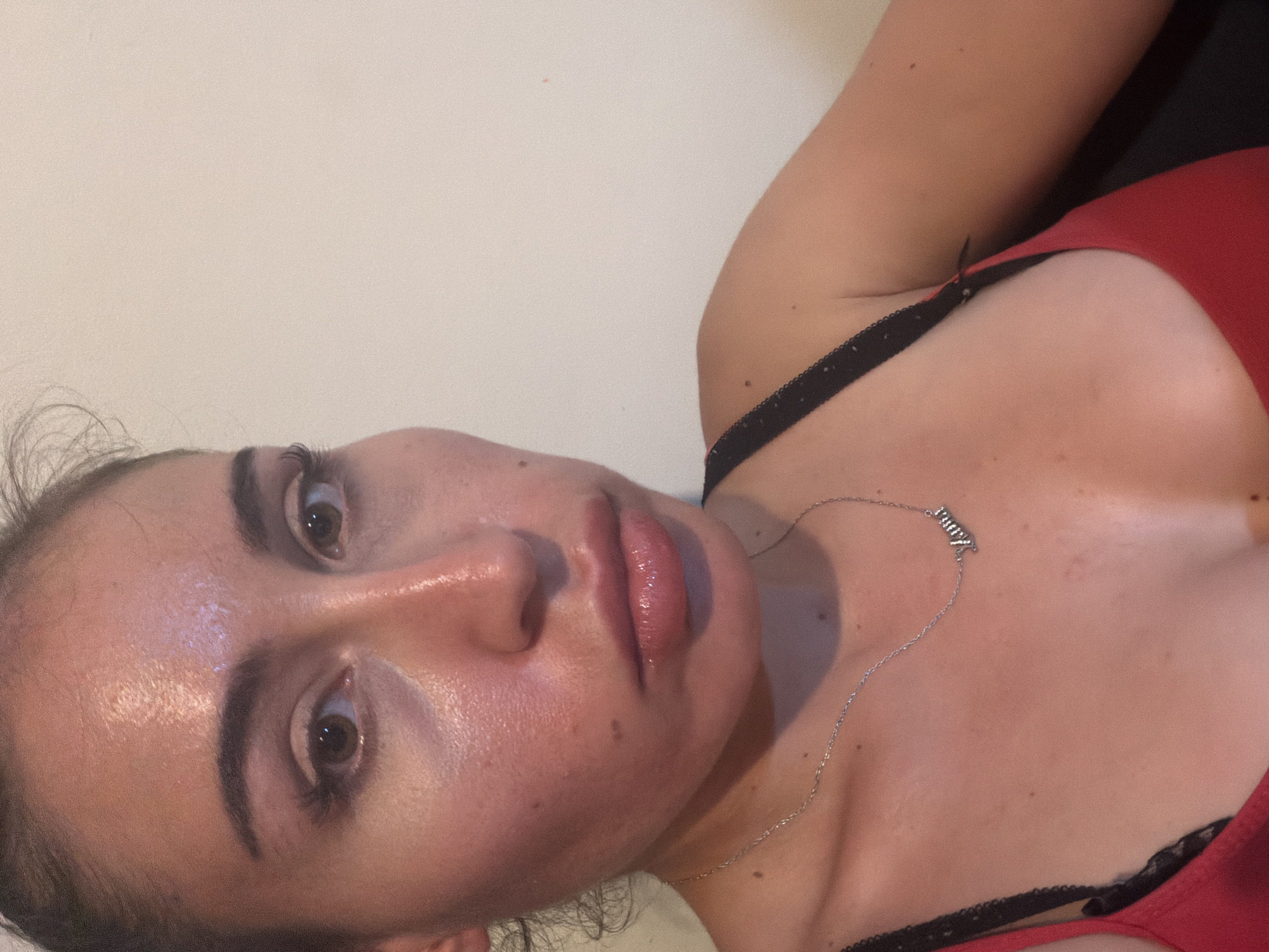 The media file contains a close-up, rotated, and low-angle shot of a woman. She is lying down, and her face is the primary focus. She has brown eyes, full lips with a glossy finish, and dark eyebrows. Her skin appears to have a dewy or oily sheen. She is wearing a red top with black lace trim and a thin silver necklace with a charm that appears to spell out "Amy" in a stylized font. The background is a plain, light-colored wall. The lighting suggests an indoor setting, possibly at night or in a room with artificial light, as there are no obvious natural light cues. The woman's expression is neutral. There are no discernible environmental cues to suggest the specific location beyond the provided context of Tersefanou, Cyprus. The image is a self-portrait or a photograph taken by someone close to the subject.
