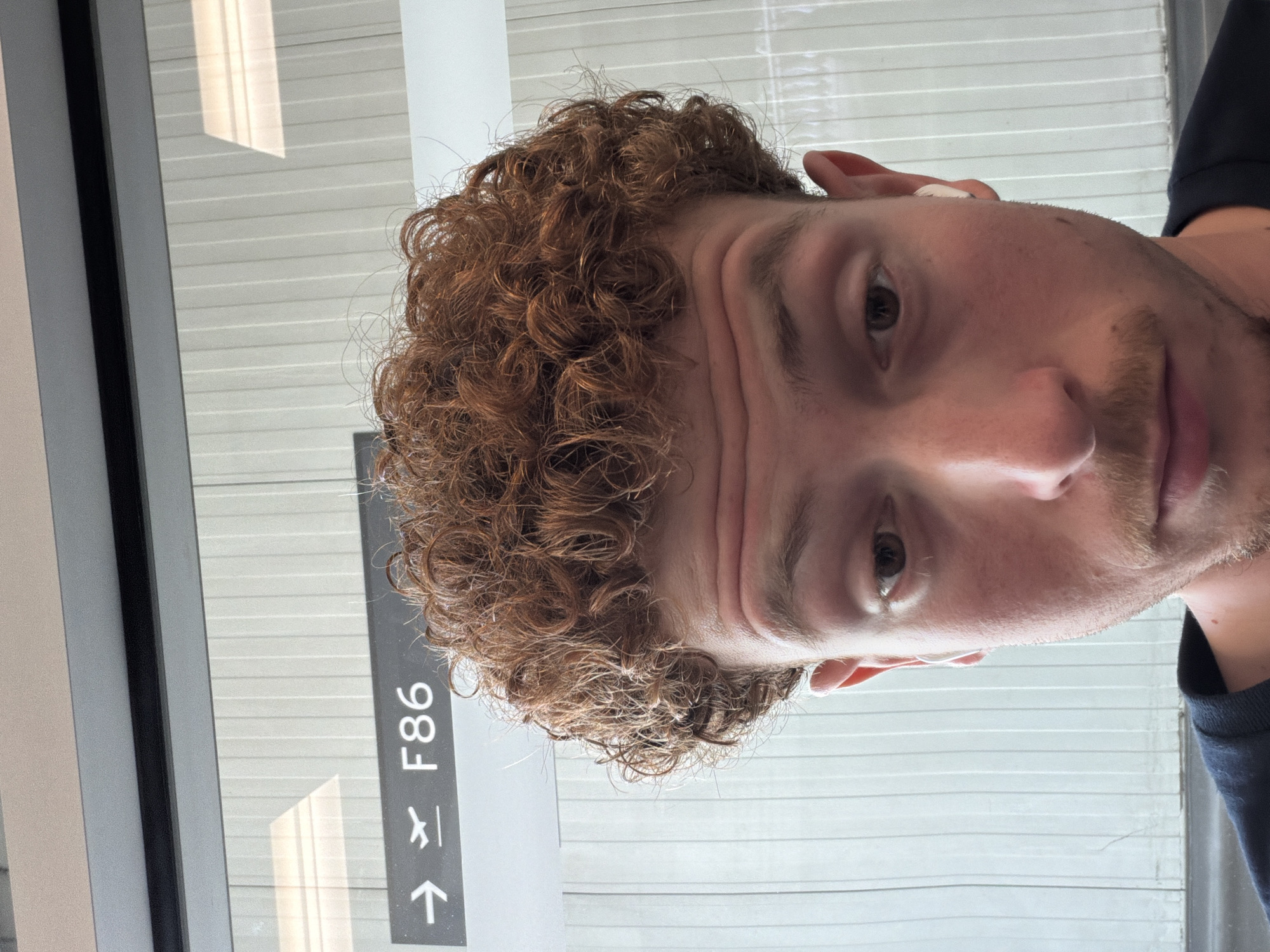 A selfie of a man with curly hair, partially visible stubble, and a light complexion, taken at an airport gate, specifically gate F86. The location is likely Toronto Pearson International Airport in Mississauga, Canada, given the context of airport gates. The background shows a window or wall with horizontal linear patterns, possibly window blinds or architectural paneling.