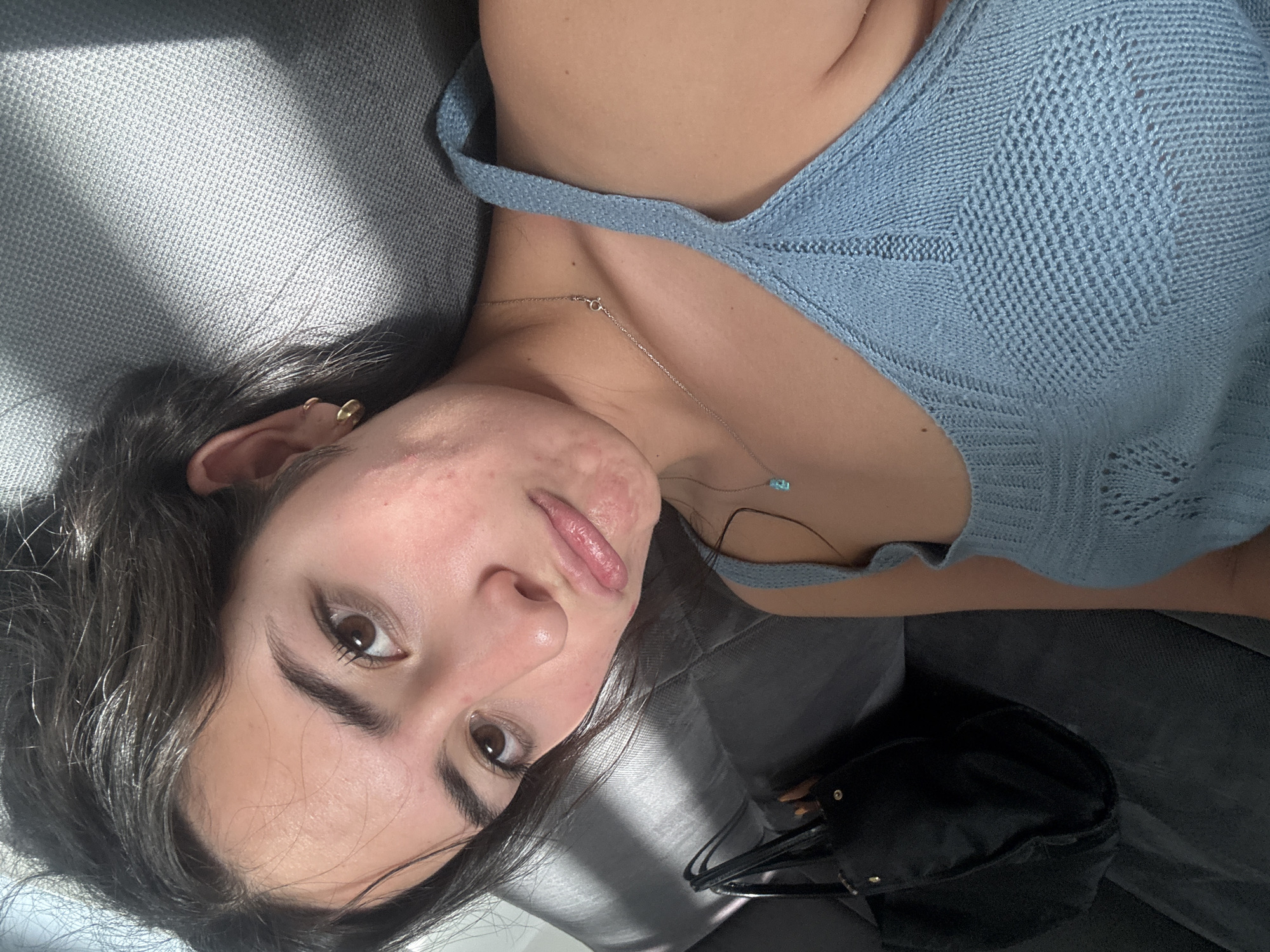 A close-up, high-angle selfie captures a young woman lying on her side, presumably on a dark gray couch or upholstered surface, indoors in San Bernardino Tlaxcalancingo, Mexico. She is looking directly at the camera with a neutral to slightly pouting expression.
The woman has dark, wavy hair framing her face, with brown eyes and well-defined eyebrows. Her left cheek shows some visible acne and texture. She is wearing a light blue, intricately knitted sleeveless top featuring a diamond-like patterned weave. Around her neck, she wears a delicate silver chain necklace with a small, light blue rectangular charm. A small gold hoop earring is visible in her right ear.
Bright, natural light, likely sunlight, streams into the room, casting strong, distinct shadows across the gray surface she is resting on and subtly illuminating parts of her face and hair, indicating it is daytime. A black bag or purse with visible metal details rests on the couch beside her. The image has an intimate and casual feel. No text is visible in the media.