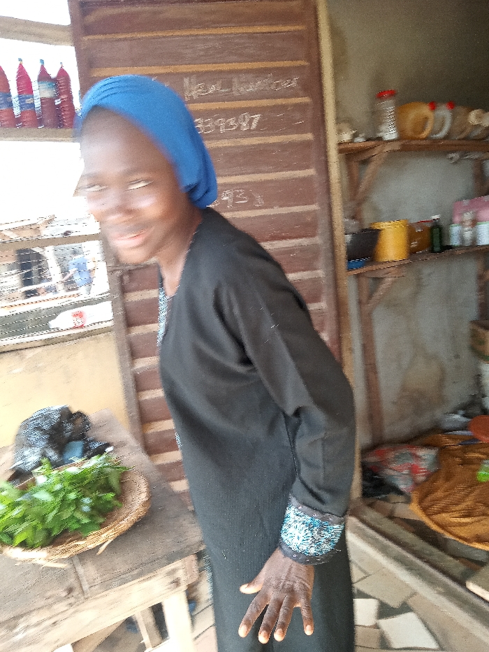 The image shows a young woman wearing a black dress and a blue headscarf. She is standing next to a wooden table with green leafy vegetables in a basket. The table is situated near a doorway, and the background shows a wooden shelf with various items on it. The woman's right hand is extended towards the camera. The location is a shop in Ota, Nigeria, with coordinates 6.691 latitude and 3.177 longitude.