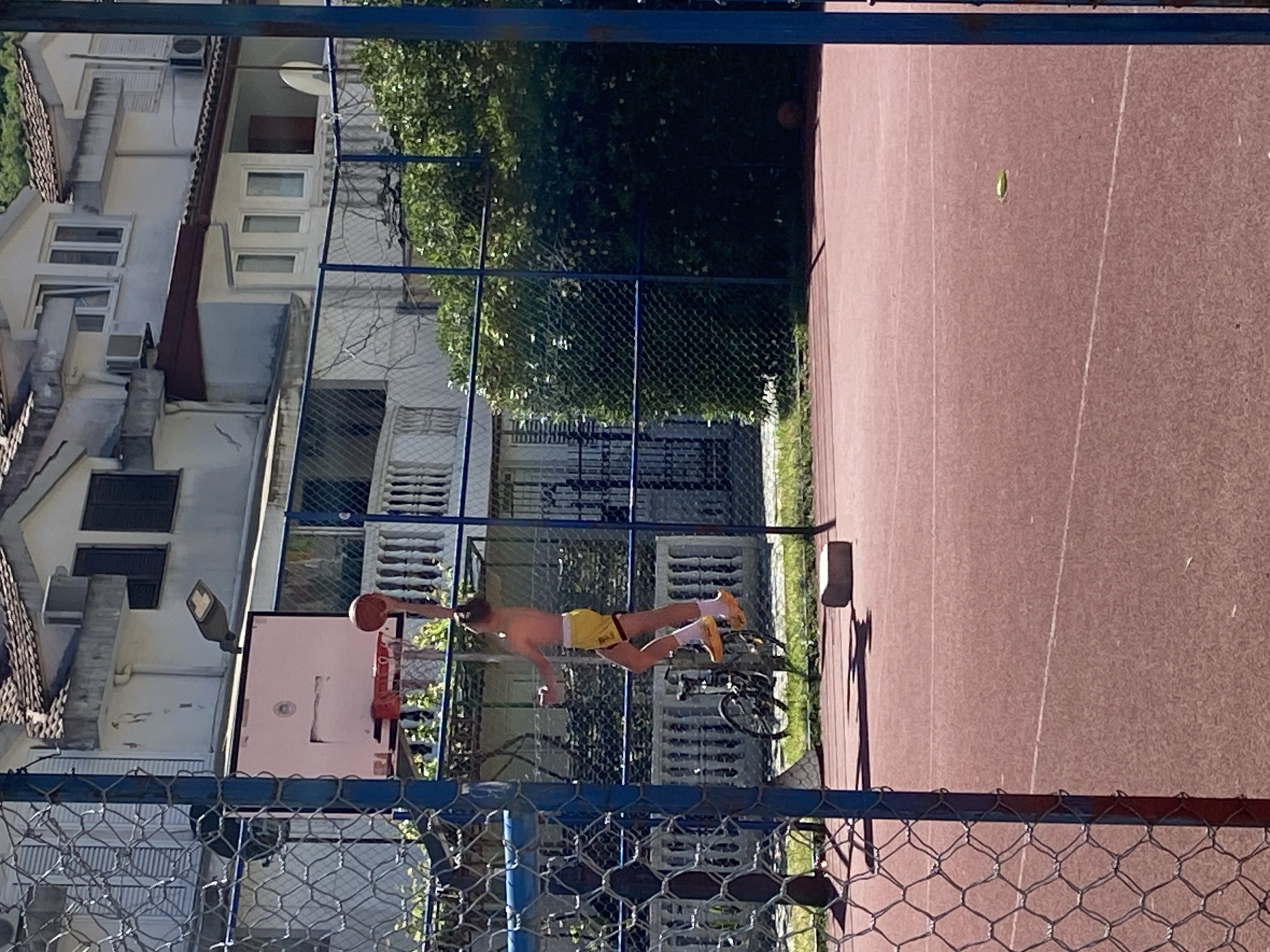 The image captures an athletic moment on an outdoor basketball court in Budva, Montenegro, under bright daylight. A young, shirtless male, wearing yellow shorts with a black 'Red Bull' branded band and light-colored shoes, is airborne, extending towards a basketball hoop to dunk an orange basketball. His body is almost horizontal in mid-air, showcasing a powerful leap.
The court itself is reddish-brown with a textured surface, and a faint white line runs across it. A small, green, elongated object, possibly a leaf, lies on the court. A bicycle is parked near the base of the basketball pole, which also supports a light fixture. A prominent chain-link fence runs across the foreground, framing the scene, and another fence is visible in the background, separating the court from surrounding areas.
Beyond the fences, several multi-story residential buildings with light-colored facades, numerous windows, balconies, and air conditioning units are visible. Their tiled roofs suggest a Mediterranean architectural style. Lush green trees and bushes fill the spaces between the buildings and behind the fences, providing a natural backdrop. The strong sunlight creates clear shadows, indicating a sunny midday or early afternoon.