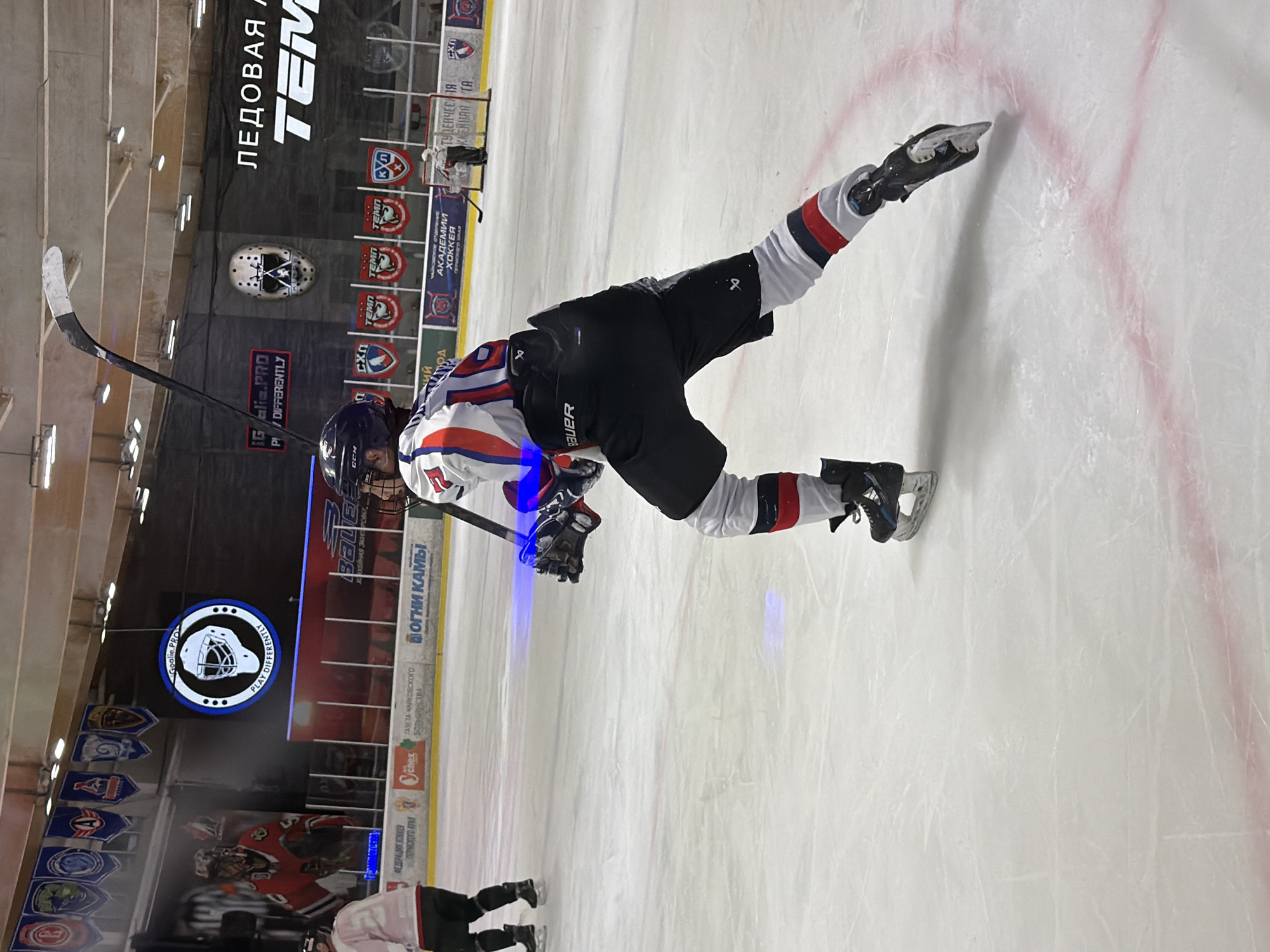 A sideways, eye-level, medium shot captures a hockey player mid-action on an ice rink. The player, wearing a blue helmet, white, blue, and red jersey, black shorts, and black and white socks with black skates, is angled towards the upper right, with one leg extended backward and the other bent. He holds a hockey stick in his gloved hands.
The background includes rink-side advertisements, a hockey goalie mask graphic, and various logos. The words "ЛЕДОВАЯ TEM" are prominent at the top. Banners and signs read "Goalie PRO PLAY DIFFERENTLY", "ХОКЕЙНАЯ ЭКИПИРОВКИ ФЕДЕРАЦИЯ ХОККЕЙ", "ВОГНИ КАМЫ ГАЗЕТА ЧАЙКОВСКОго БОЛЬЦИНСТВА", and "IGalia.PRO P/ DIFFERENTLY", identifying the location as Chaikovsky, Russia. The ice surface shows skate marks and red lines. Another player is partially visible in the lower left.