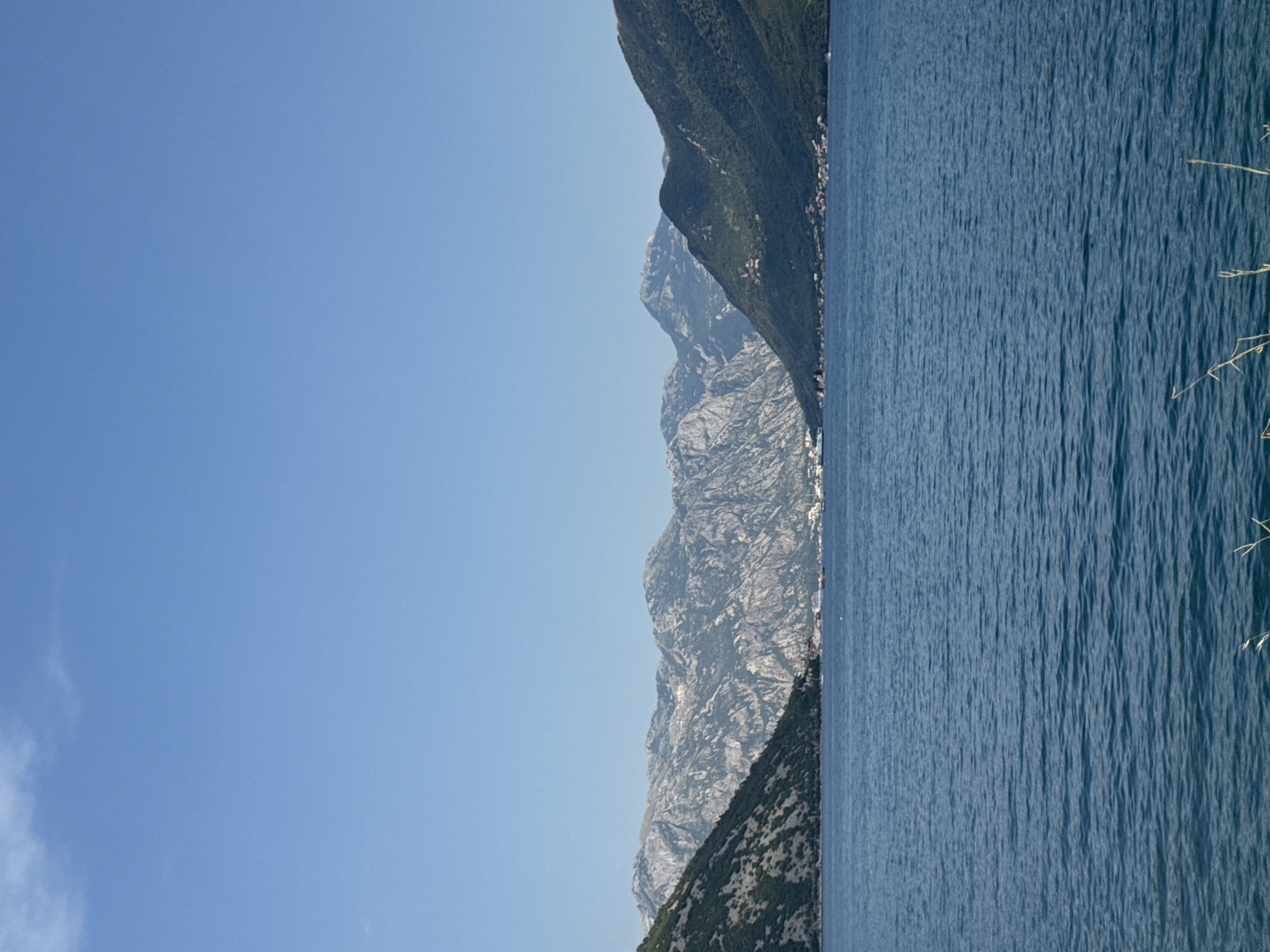 The image depicts a serene body of water, likely a bay or fjord, with a clear blue sky overhead. In the background, majestic mountains rise, their rocky slopes covered in sparse vegetation and some patches of bare rock. The mountains form a dramatic backdrop to the tranquil water. Along the base of the mountains, a settlement or town is visible, characterized by small buildings and a hint of urban development nestled into the landscape. The water is calm, with gentle ripples reflecting the blue of the sky and the surrounding environment. The overall scene conveys a sense of natural beauty and peacefulness, characteristic of coastal mountain regions. This location is identified as Lipci, Montenegro.