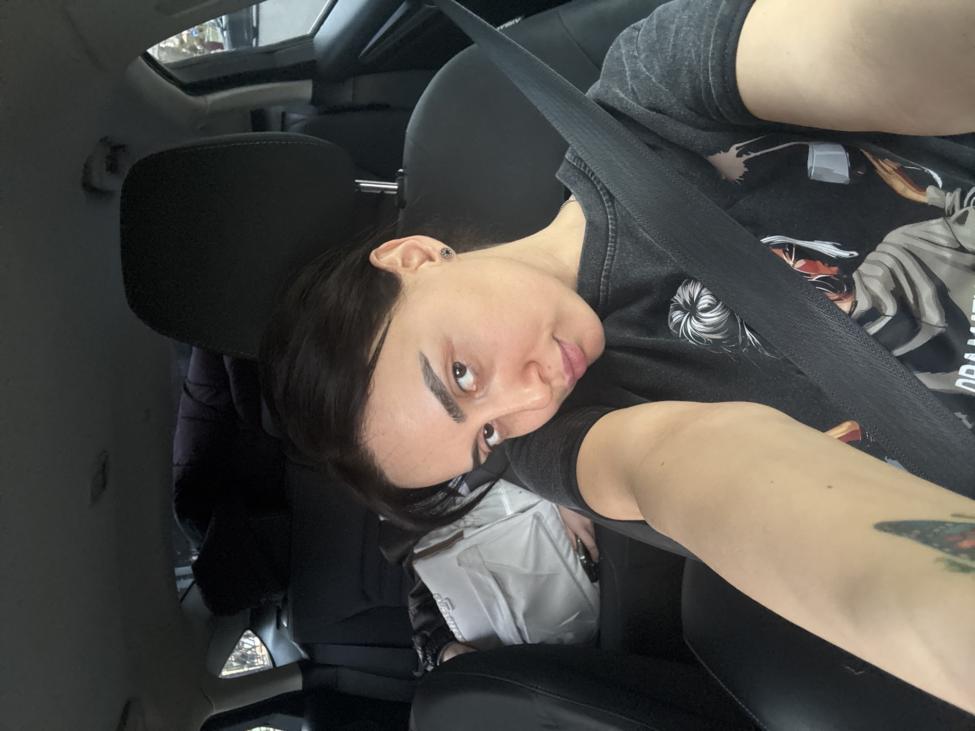A selfie taken inside a car. A person with dark hair, wearing a gray t-shirt and a seatbelt, is looking into the camera. The interior of the car is visible, including seats and a sunroof. There is a white bag on the passenger seat. The exact location in Tbilisi, Georgia cannot be determined from this image alone.