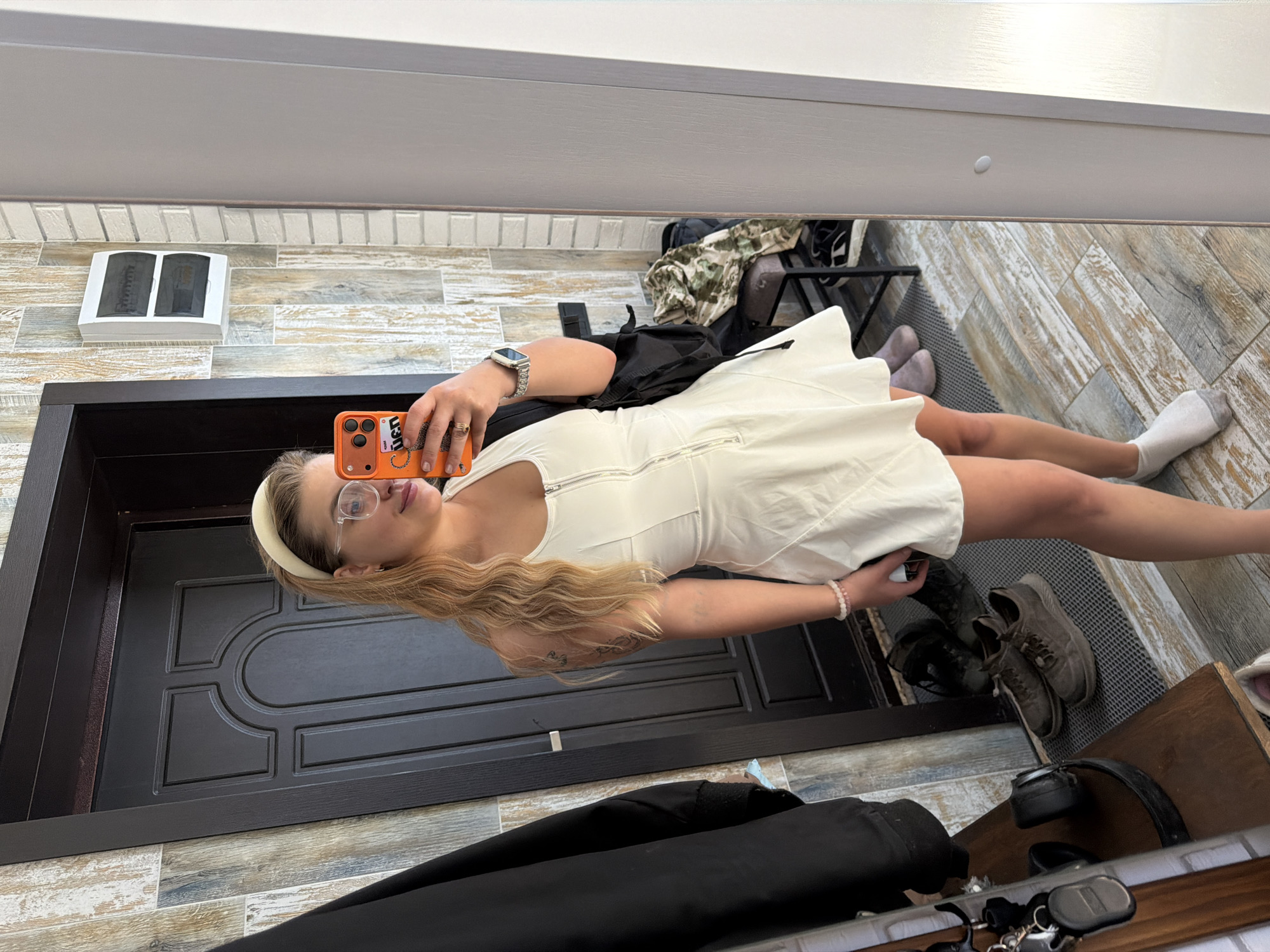 A woman is taking a selfie in front of a mirror. She is wearing a white dress and glasses, with a headband on her head. She is holding an orange phone in her hand.
The location is suggested to be in Большая Шильна, Russia.
The background features a wooden door, wood-look tiled walls, and a glimpse of the floor. There are shoes, bags and other items visible near the bottom and top right of the frame.