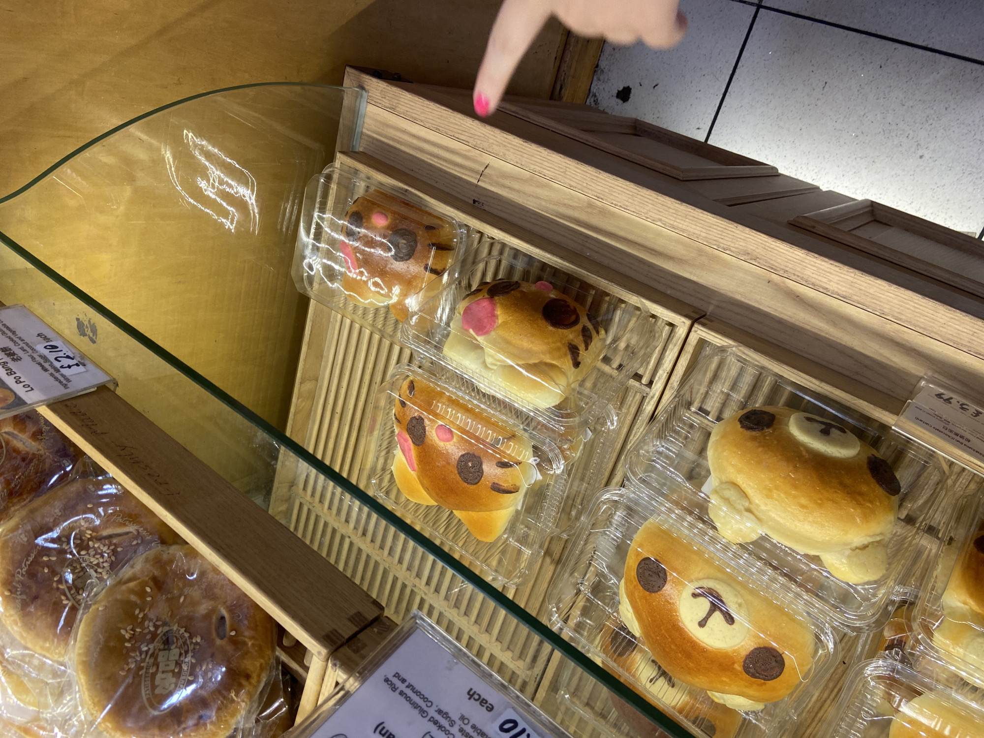 This image shows a display case in what appears to be a bakery or shop, possibly in London, United Kingdom. The display features different types of sweet buns, including "Lo Po Bang" pastry and "Rilakkuma Bun". The pastries are enclosed in clear plastic containers within wooden shelves. A hand with pink nail polish is pointing at one of the containers in the image. Pricing is visible on labels near the buns, in both GBP and another language. The pastries on display have decorative features, resembling bear faces and other animal-like designs.