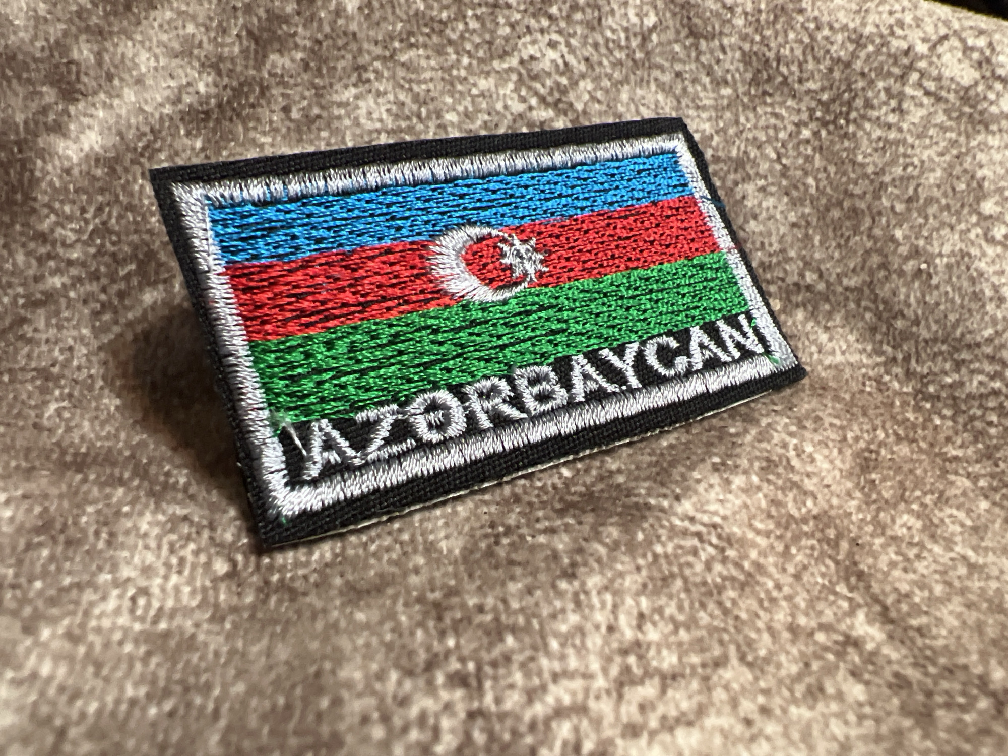A close-up, top-down shot shows an embroidered patch of the flag of Azerbaijan on a textured, brown fabric. The patch is rectangular and features horizontal stripes of blue, red, and green, with a white crescent moon and star in the center of the red stripe. The word "AZERBAYCAN" is embroidered in white block letters along the bottom of the flag. The background fabric appears to be a fuzzy, woven material, possibly a blanket or upholstery. The lighting is dim, casting soft shadows. The specific location within Masazir, Azerbaijan, is not discernible from the image.