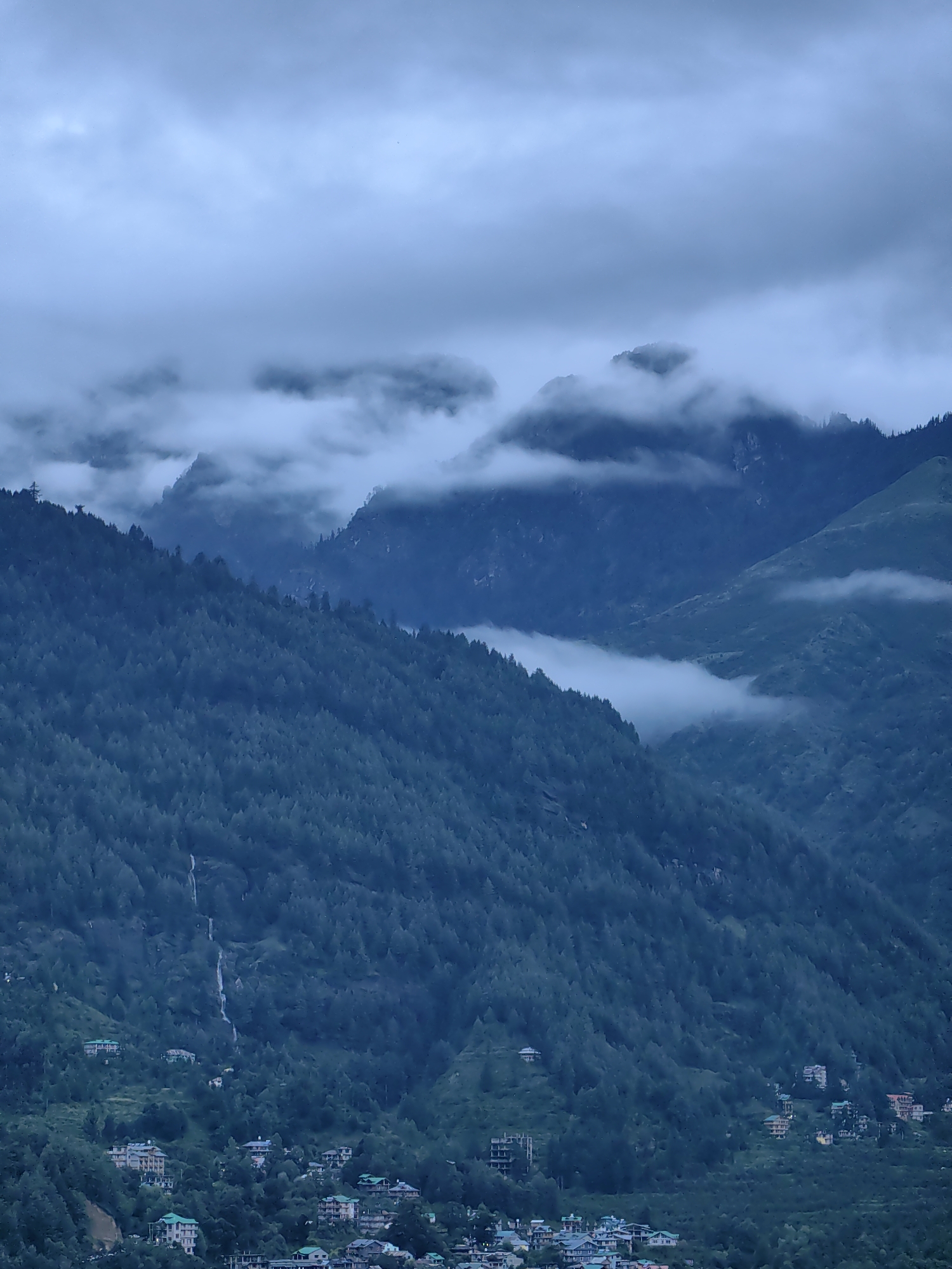 The image depicts a panoramic view of the mountainous landscape surrounding Vashisht, India, under a cloudy sky. The foreground features a dense forest covering the steep slopes of the mountains. Scattered throughout the hillside are numerous buildings, indicating a settlement nestled within the natural environment. Some of these structures appear to be hotels or guesthouses, characterized by their multi-story designs and pitched roofs, many of which are green. A thin waterfall is visible cascading down the forested slope. The background is dominated by more mountains partially obscured by mist and low-hanging clouds. The overall atmosphere is serene and natural, with a hint of human habitation integrated into the landscape.