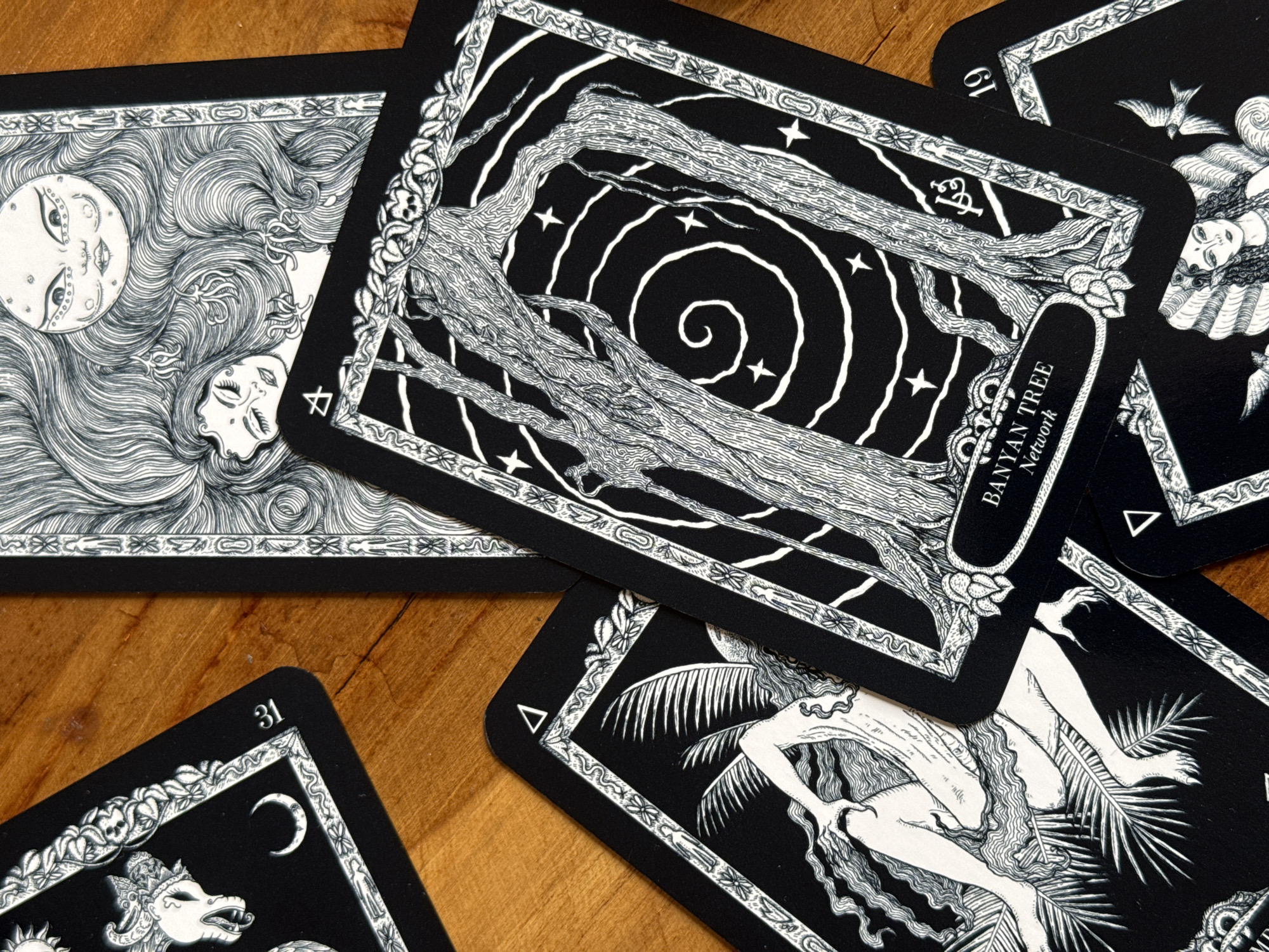 The image shows a collection of tarot-style cards spread on a wooden surface. The cards are black and white with intricate illustrations. One prominent card features a Banyan Tree with spiraling white lines and stars in the background; the card reads "BANYAN TREE Network." Another card displays a face with long flowing hair, accompanied by a moon. A third card partially visible showcases a figure with outstretched arms and palm fronds. Numbers appear in the upper left corner of some cards, such as "31." The setting is not identifiable as North Kuta, Indonesia.