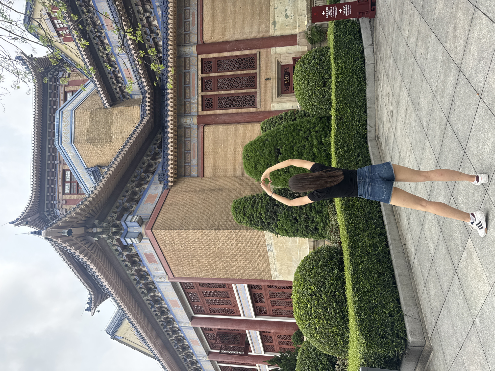 A full shot of a person standing on a grey tiled surface in front of a traditional Chinese building and several rows of neatly trimmed shrubbery. The building is made of red brick, with several windows. The person is seen from behind, wearing a black t-shirt and blue denim shorts. The person is holding their arms up, forming a circular shape. The image is taken during the day, likely in the Yuexiu District of China.