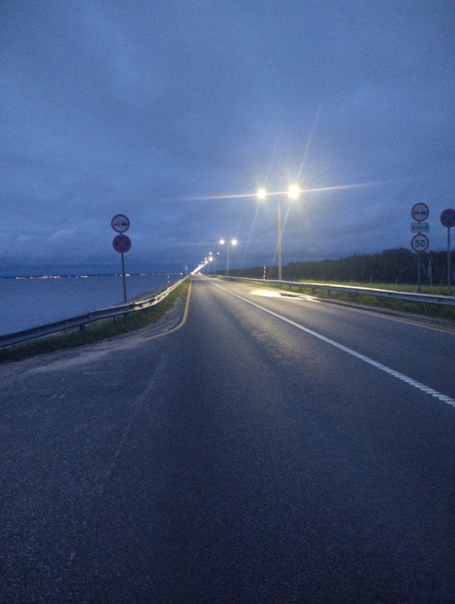 A highway runs alongside a body of water at dusk or dawn. The sky is a deep blue with clouds. Streetlights illuminate the road, creating bright glare. On the left side of the road, there are several traffic signs: a sign with two red circles, another sign with a red circle and a diagonal bar, and a circular sign with the number 50 inside a red circle. The body of water on the left has lights visible in the distance, suggesting it's a town or city. Trees line the right side of the road. The road surface is asphalt with white lane markings. There are no people or vehicles visible in the image. The scene is in Zavolzhye, Russia.