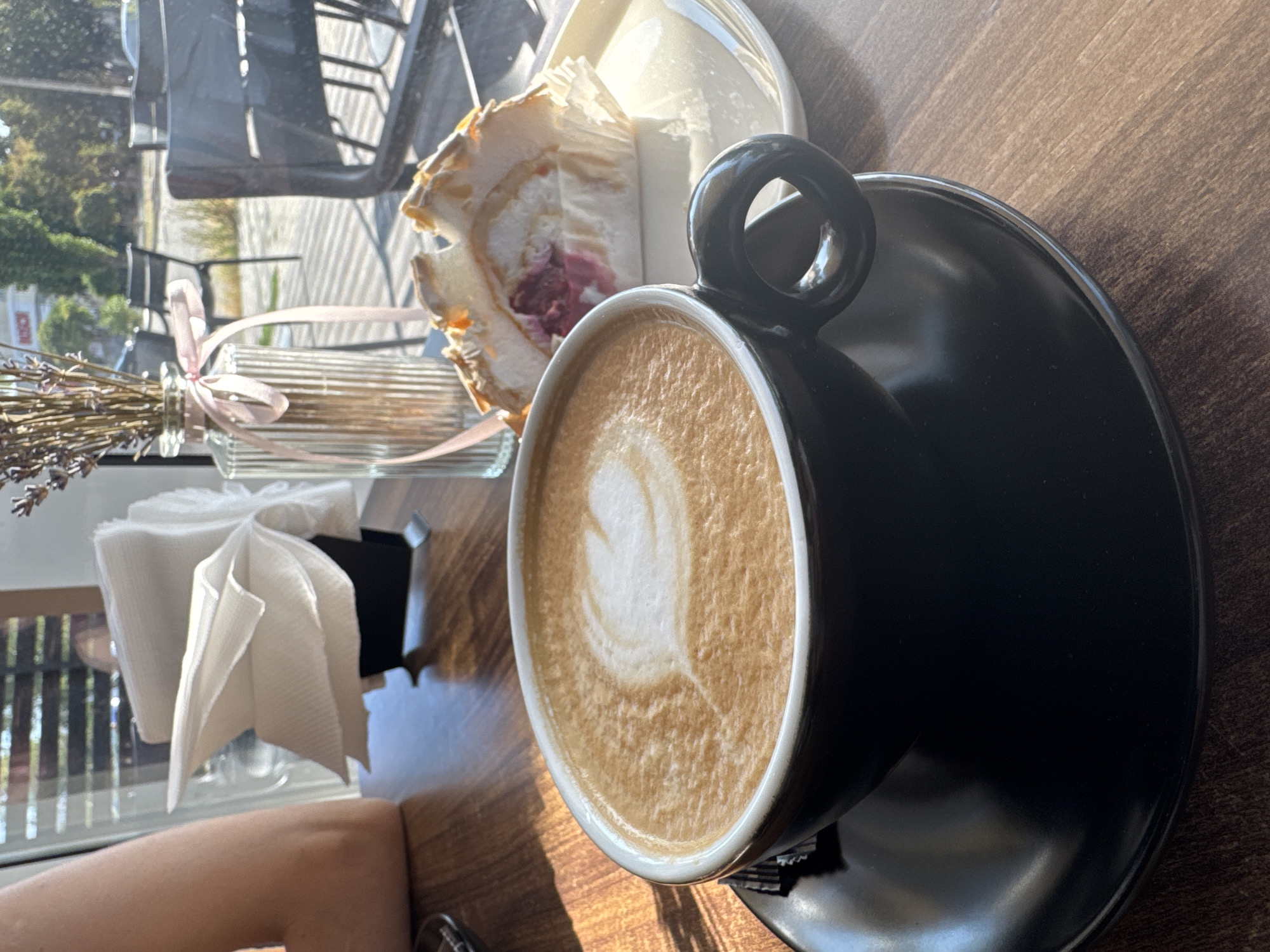 This is a close-up, eye-level, slightly angled shot of a cappuccino with latte art in the shape of a heart, served in a black mug and saucer on a wooden table. Beside the coffee, on a white saucer, is a slice of meringue roll with a pink filling and a meringue topping adorned with slivered almonds. In the background, a glass vase with dried lavender tied with a pink ribbon is visible, along with a stack of white napkins in a black holder. Through a window behind these items, outdoor seating with black chairs and tables, trees, and buildings are discernible, suggesting an outdoor cafe setting in Bălți, Moldova. Sunlight streams in from the left, casting shadows on the table. The arm of a person is partially visible on the far left of the frame.