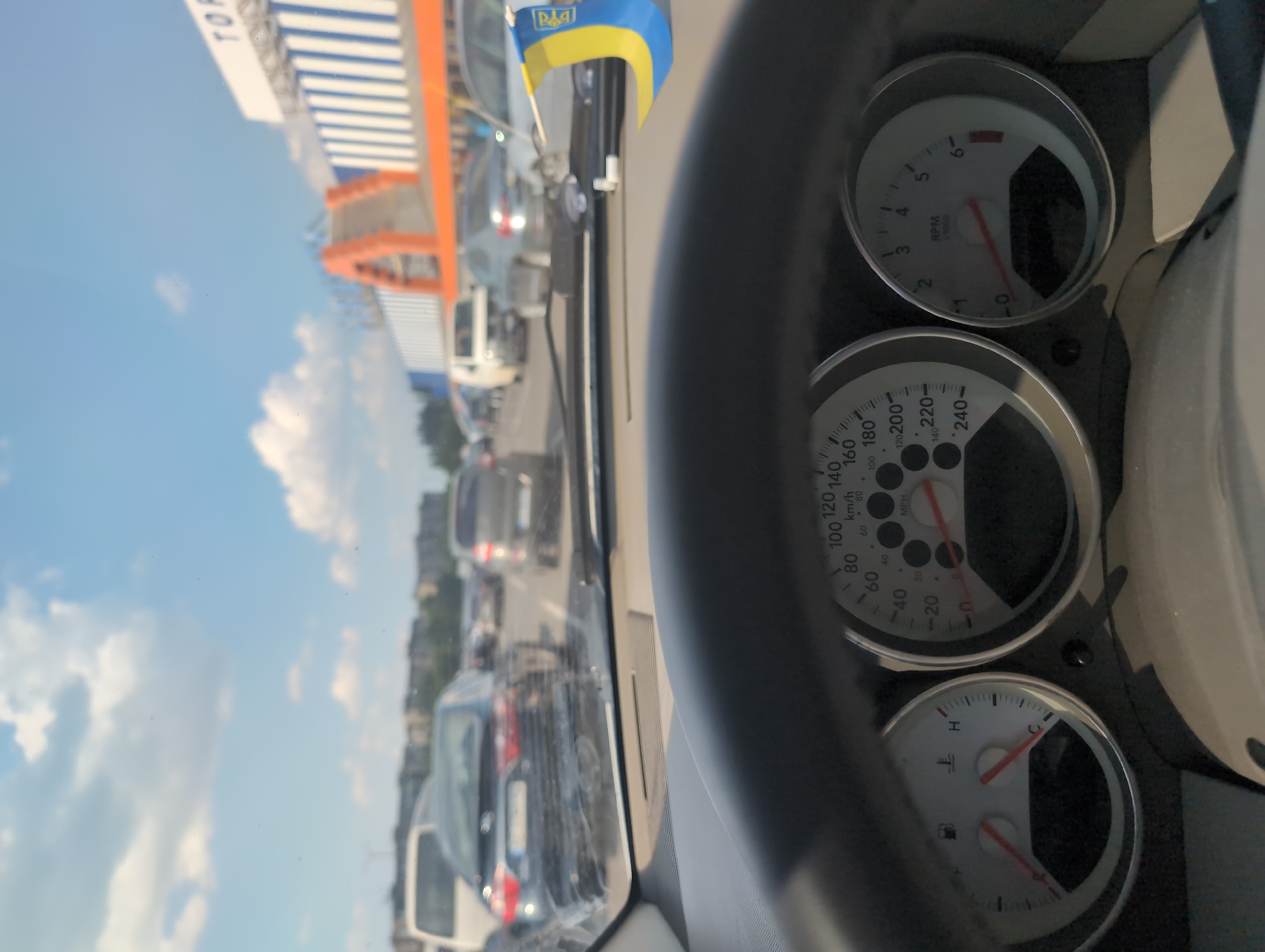 This image appears to be taken from inside a car, looking forward through the windshield. The driver's perspective reveals a dashboard with two prominent gauges: a speedometer that can display speeds up to 240 km/h and 240 MPH, and a tachometer indicating RPMs up to 6000. A smaller gauge, likely for fuel or temperature, is also visible. A small Ukrainian flag is attached to the car's interior.
Outside the car, a traffic jam is visible on a sunny day. The sky is blue with scattered white clouds. The road ahead is lined with other vehicles, suggesting a congested urban or highway environment. In the background, a structure with orange and white stripes, possibly part of a building or a roadside attraction, is visible. The overall scene suggests a moment of being stuck in traffic on a clear day. The visible text includes "RPM x 1000" and speed indicators on the gauges, as well as what appears to be a partial sign with "TO" on it. The location context provided is Kryvyi Rih, Ukraine.