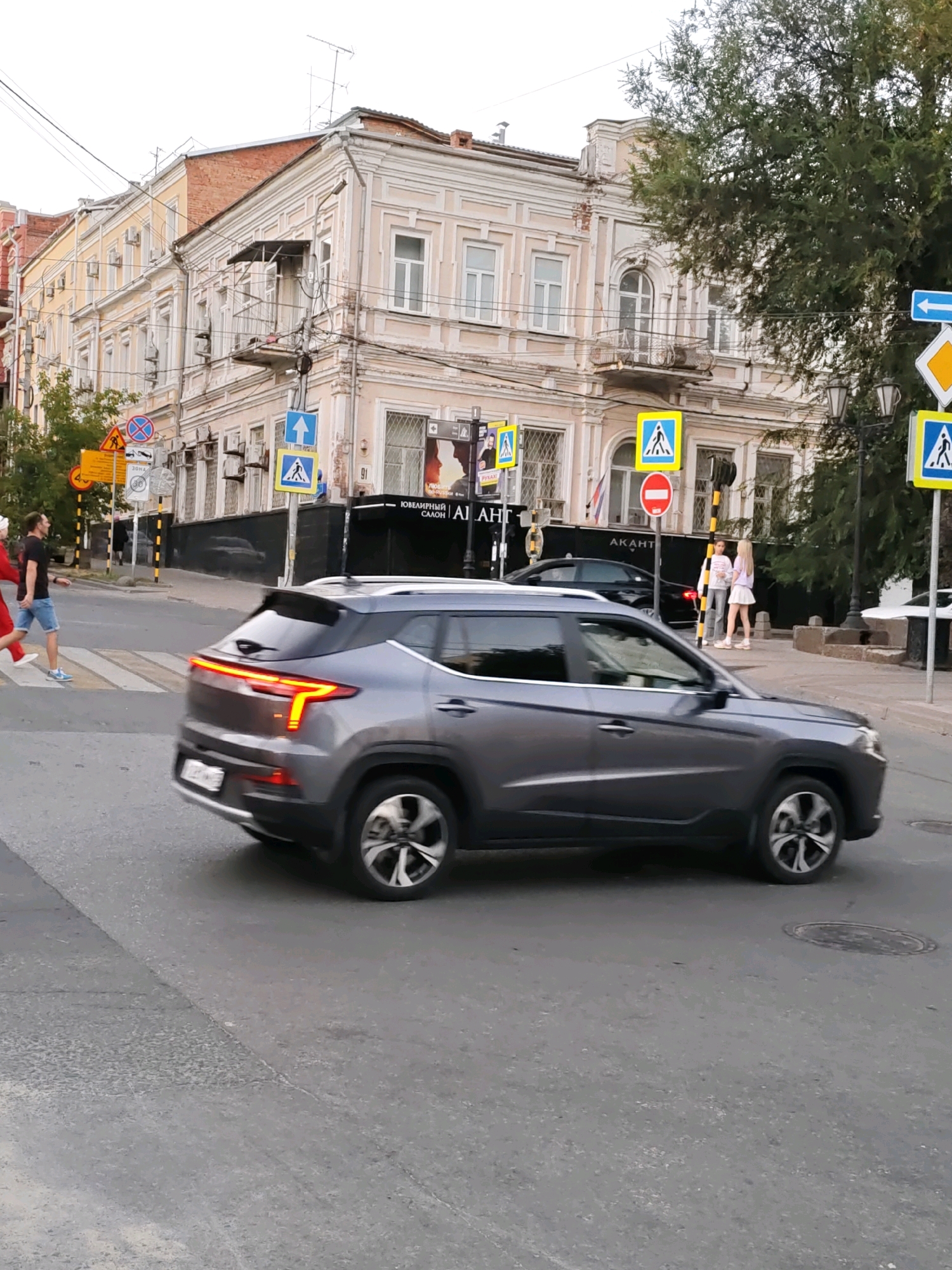 A gray SUV is captured in motion, crossing a pedestrian street in the city of Rostov-on-Don, Russia. The vehicle is angled from the side, with its rear lights illuminated. In the background, a multi-story building with ornate architecture stands, featuring large windows, balconies, and decorative moldings. Pedestrian crossing signs, some displaying a blue square with a white pedestrian symbol, are visible on the street. A traffic sign with a red circle and horizontal white bar indicates "no entry." Two young women, one in a light-colored skirt and the other in a white top and skirt, are standing on the sidewalk, conversing. To the left, a man in a black t-shirt and blue shorts walks across the street, while another person in a red outfit is also crossing. A green tree with dense foliage partially obscures the building on the right. The sky is overcast.