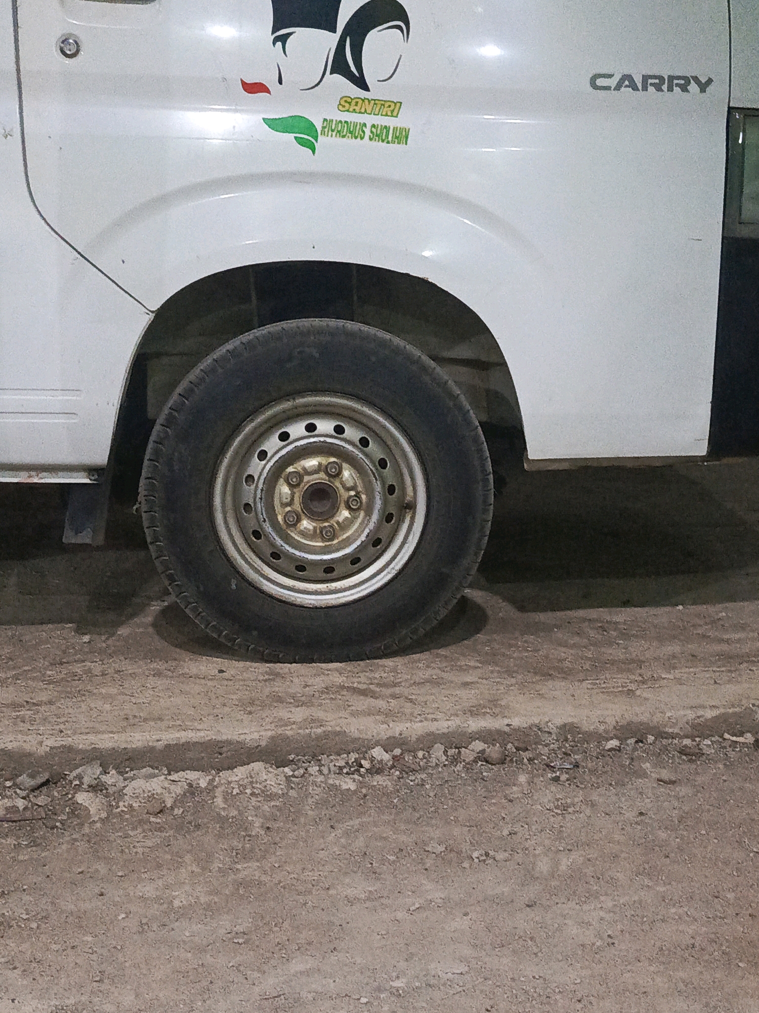 The image shows the rear passenger-side wheel and lower body panel of a white Suzuki Carry utility vehicle. A decal on the vehicle's body depicts two stylized human figures wearing head coverings, with text beneath that reads "SANTRI RIYDHUS SHOLIHAIN." The word "CARRY" is also visible in silver lettering on the vehicle's panel. The vehicle is parked on a dirt and gravel surface. The background is dark, suggesting it is nighttime or an indoor location with poor lighting. There is no clear indication of the specific location within Megang Sakti V, Indonesia, as no landmarks are visible.