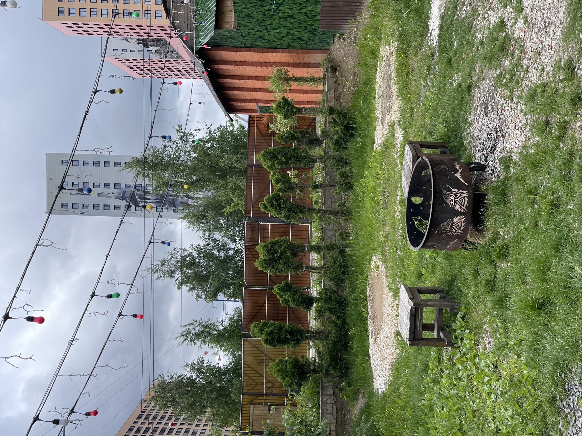 The image shows an outdoor area in Kommunarka, Russia. In the foreground is a metallic fire pit with decorative cutouts and a wooden bench. The ground is a mix of grass and gravel. A line of small trees separates the foreground from the background. Behind the trees is a fence and several buildings, including a brick structure and tall residential buildings with visible windows. Overhead, strings of colorful lights are strung across the area. The sky is overcast.