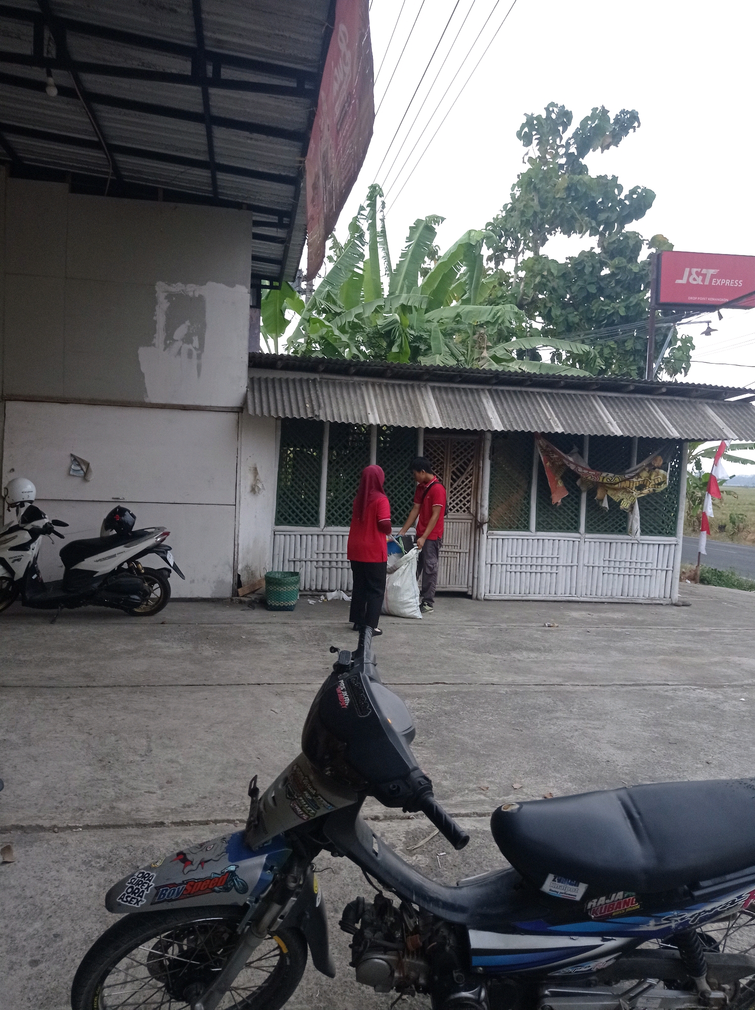 In Jetis, Indonesia, a scene unfolds outside a small, open-fronted building with a corrugated metal roof and a bamboo-slatted facade. Two individuals are present. A person wearing a red shirt and headscarf stands to the left, holding a large white bag. To their right, a person in a red shirt and dark pants is interacting with the bag, appearing to be placing items into it.
In front of the building, two motorcycles are parked on a concrete surface. The motorcycle on the left is white with a black seat and a white helmet resting on it, along with another helmet on the handlebars. The motorcycle on the right is primarily black and blue, with visible stickers and aftermarket parts, its front wheel and engine area are prominently featured in the foreground.
Behind the building, lush green banana trees and other foliage are visible, suggesting a semi-rural or tropical environment. A red sign with white lettering, reading "J&T EXPRESS," is mounted on a pole to the right of the building. The sky above is overcast, and a road is visible in the background.