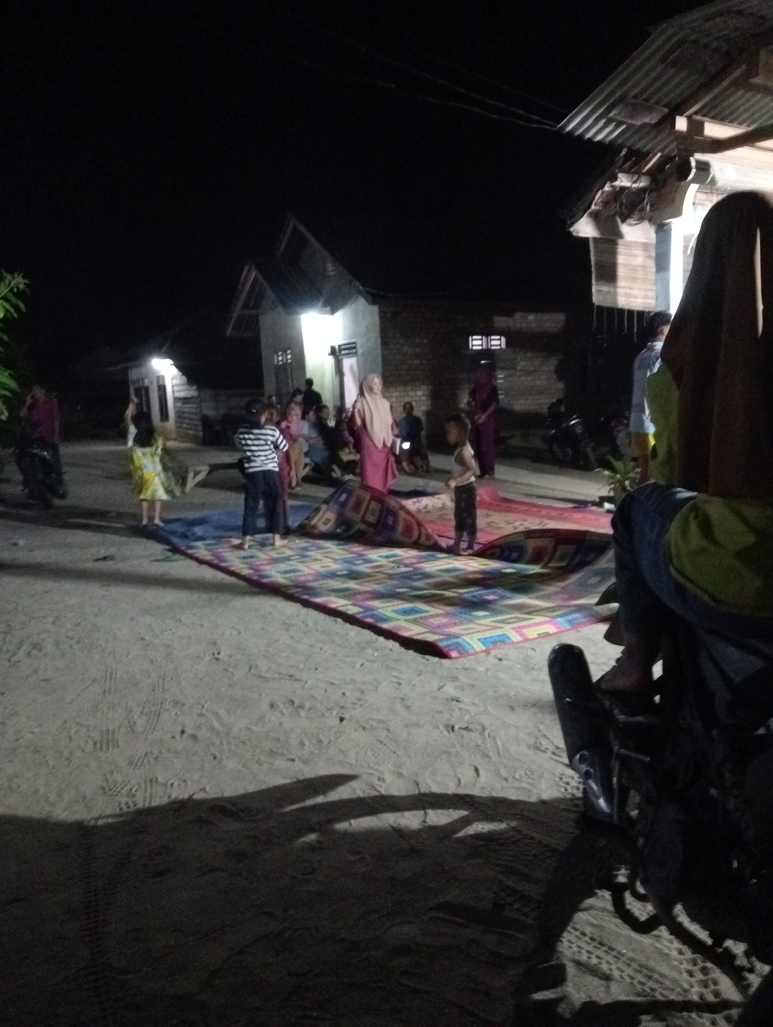 The image depicts an outdoor gathering at night in Rasuan Baru, Indonesia. Several individuals are present, including children and adults. A colorful mat is spread on the sandy ground, and some people are standing or sitting near it. In the background, there are houses with illuminated windows. The scene suggests a communal or family event taking place in a residential area after dark.