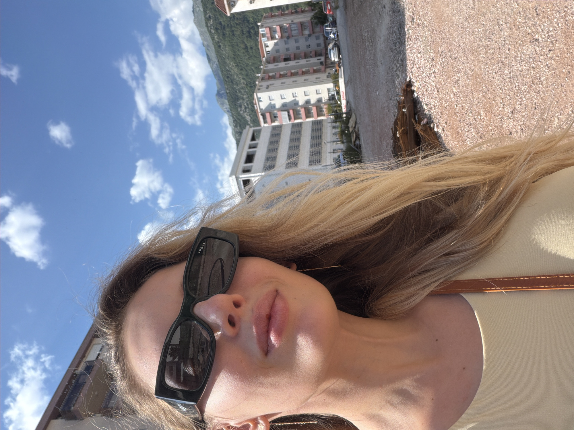 A close-up, selfie-style shot captures a blonde woman wearing dark, rectangular sunglasses and a light yellow top. She has delicate threader earrings in her ears. The background suggests a bright, sunny day with a blue sky dotted with clouds. In the mid-ground, a cluster of modern, multi-story apartment buildings is visible, some with balconies. The foreground and immediate background feature a patch of dirt and gravel, with some scattered debris, indicating a possible construction site or undeveloped area. Mountains covered in green foliage rise in the far distance, providing a scenic backdrop. This scene is consistent with an outdoor location in Budva, Montenegro, known for its coastal development and mountainous terrain.