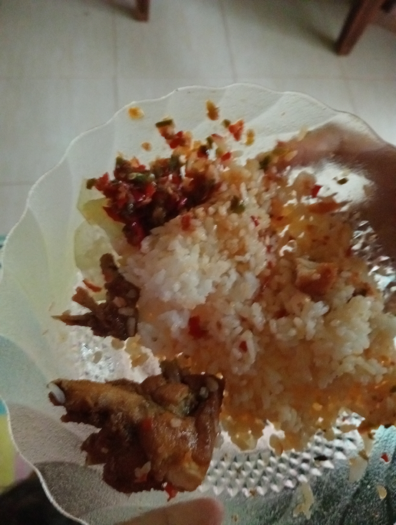 The image shows a plate of rice, fried chicken, and a chili condiment, held by a person's hand. The food is served on a transparent, scalloped-edge plate, which is resting on a textured surface, possibly a placemat or table. The background consists of a light-colored tiled floor and blurred furniture, suggesting an indoor setting. The lighting is somewhat dim, with a slight glare on the plate and food. The scene appears to be a casual meal setting in a home. The location is Campago Barat, Indonesia.