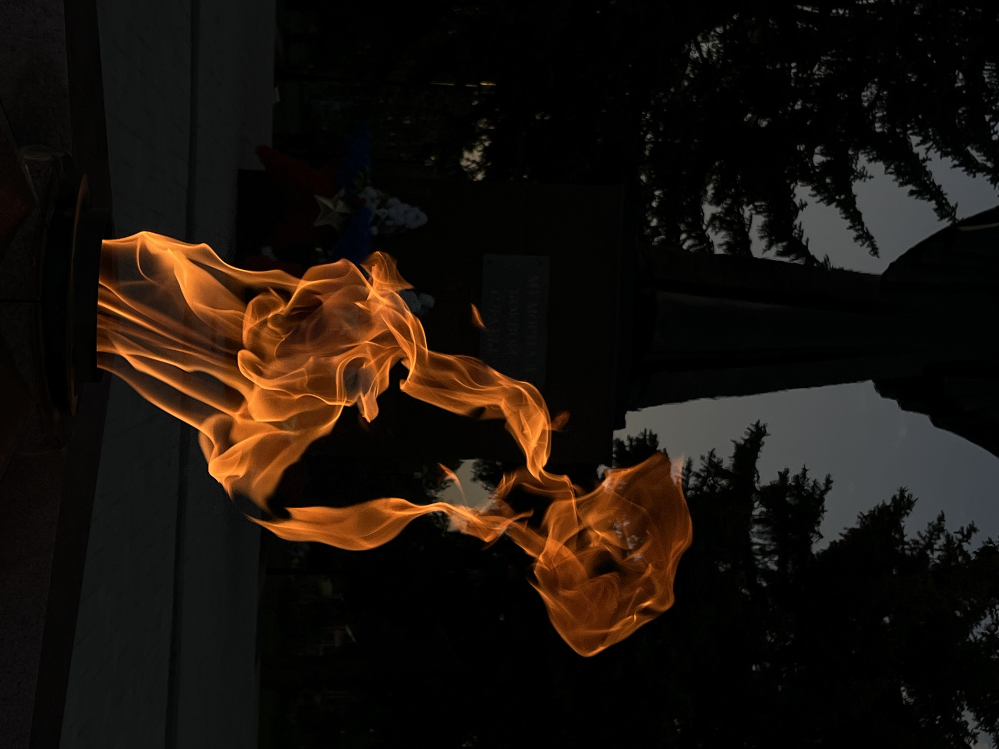 The image shows a close-up of a flame emanating from a dark, metallic structure, likely a memorial or eternal flame installation. The flame is a vibrant orange, contrasting sharply with the dark background. Behind the structure, there is a partially visible monument or headstone, with some flowers and what appears to be a star emblem. Dark silhouettes of trees are visible against a grey sky, likely indicating twilight or nighttime. The setting seems to be a memorial site or cemetery, with the eternal flame serving as a central element. Location information suggests this could be in Novopokrovskaya, Russia. The image is rotated counterclockwise.