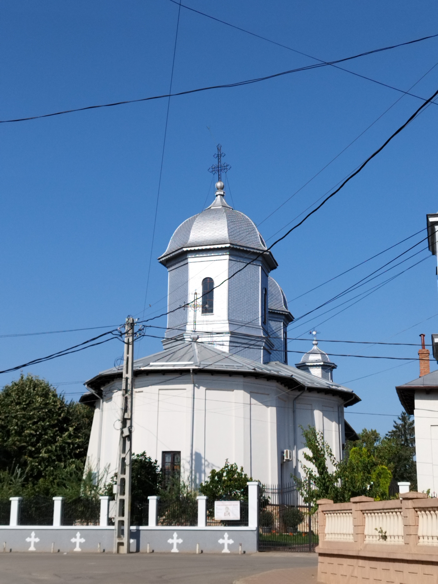 This is a daytime, eye-level, outdoor, full-shot photograph of a white church with a silver dome in Băleni-Români, Romania. The church has a circular base and a pointed roof, with a smaller dome visible on top. The main dome is topped with a cross. The church is situated in a residential area, with trees and fences visible in the foreground. The sky is clear and blue. Several electrical wires are visible in the sky, crossing the frame. There are no people visible in the image. The overall impression is one of a quiet, serene day in a small Romanian town.