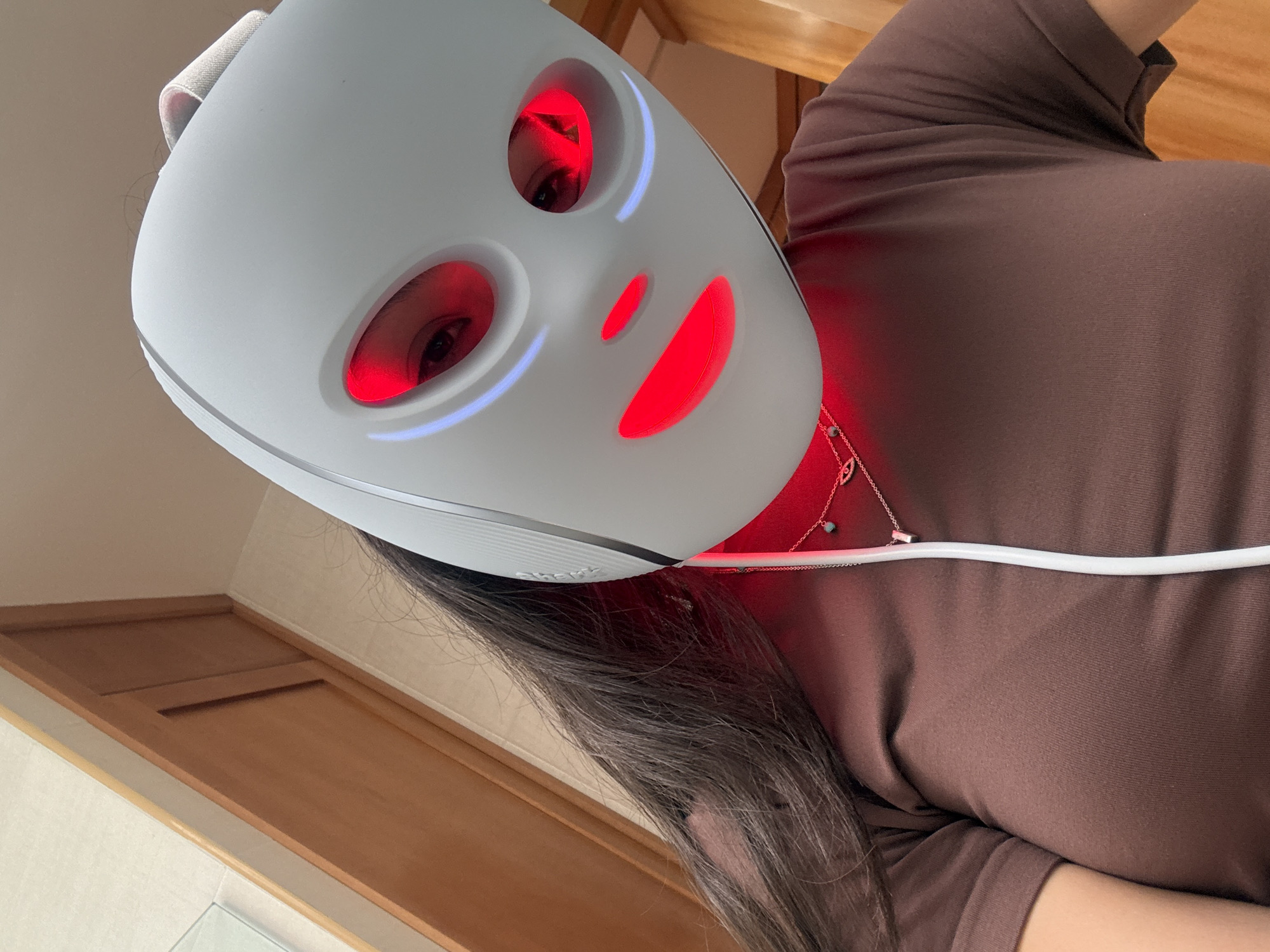 A person is wearing a white LED facial mask with red light therapy on the eye, nose, and mouth areas, and blue light around the eye areas. The mask is connected to a white cord. The person is wearing a brown long-sleeved shirt and a silver necklace. The background includes a wooden door and a wall. The location is San Bernardino Tlaxcalancingo, Mexico.
