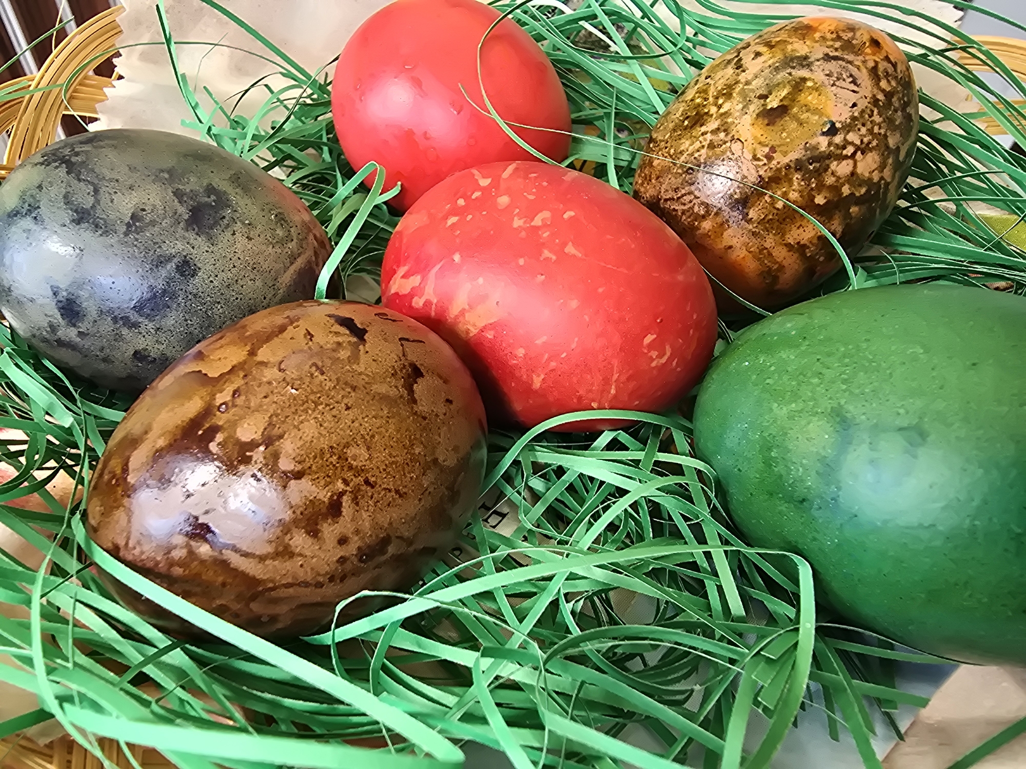 A high-angle, close-up shot depicts six decorated Easter eggs nestled among green paper Easter grass in a woven basket. The eggs are colored red (2), brown, dark brown, green, and a brownish-yellow egg with spots. The eggs are glossy, suggesting they might be boiled eggs. The basket's wickerwork is visible at the edge of the frame, and a light, waxy paper lines the basket beneath the grass. The scene is well-lit, emphasizing the colors and textures of the eggs and grass. The location is Unknown, Unknown.