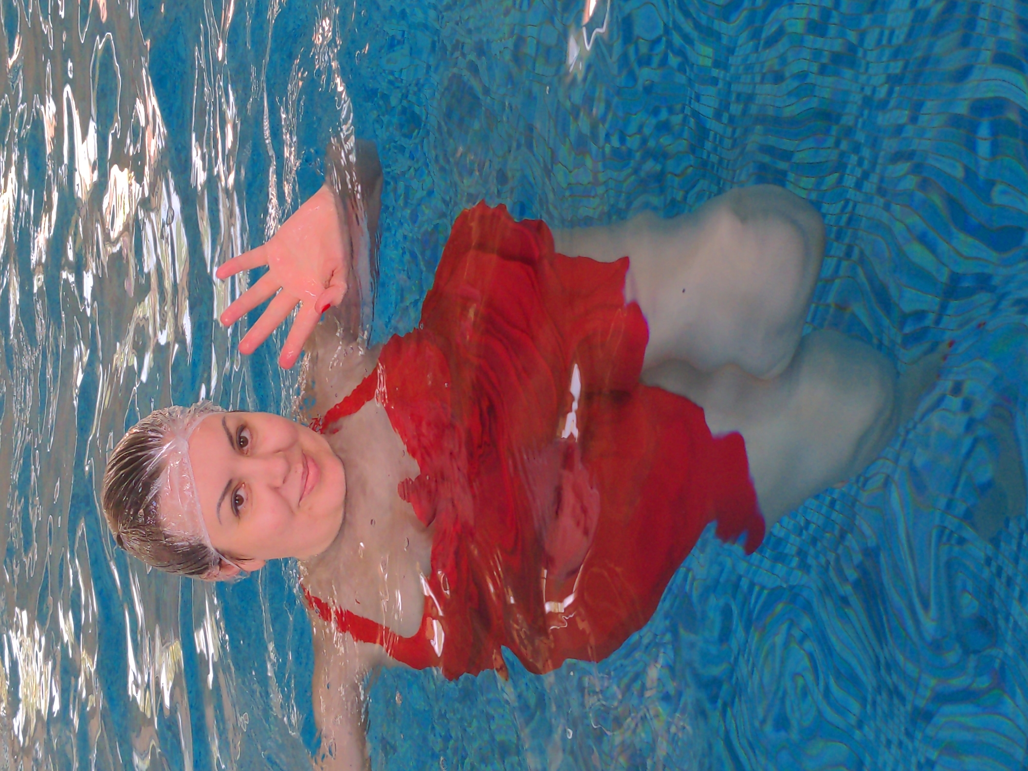 In Holubyne, Ukraine, a woman wearing a red bathing suit and a clear shower cap is submerged in a swimming pool. She is looking towards the camera and smiling. Her right hand is raised out of the water, with her fingers spread apart. The pool water is clear with blue tiled bottom visible. The lighting suggests an indoor setting, possibly a spa or water therapy center. The image captures a moment of relaxation and enjoyment.