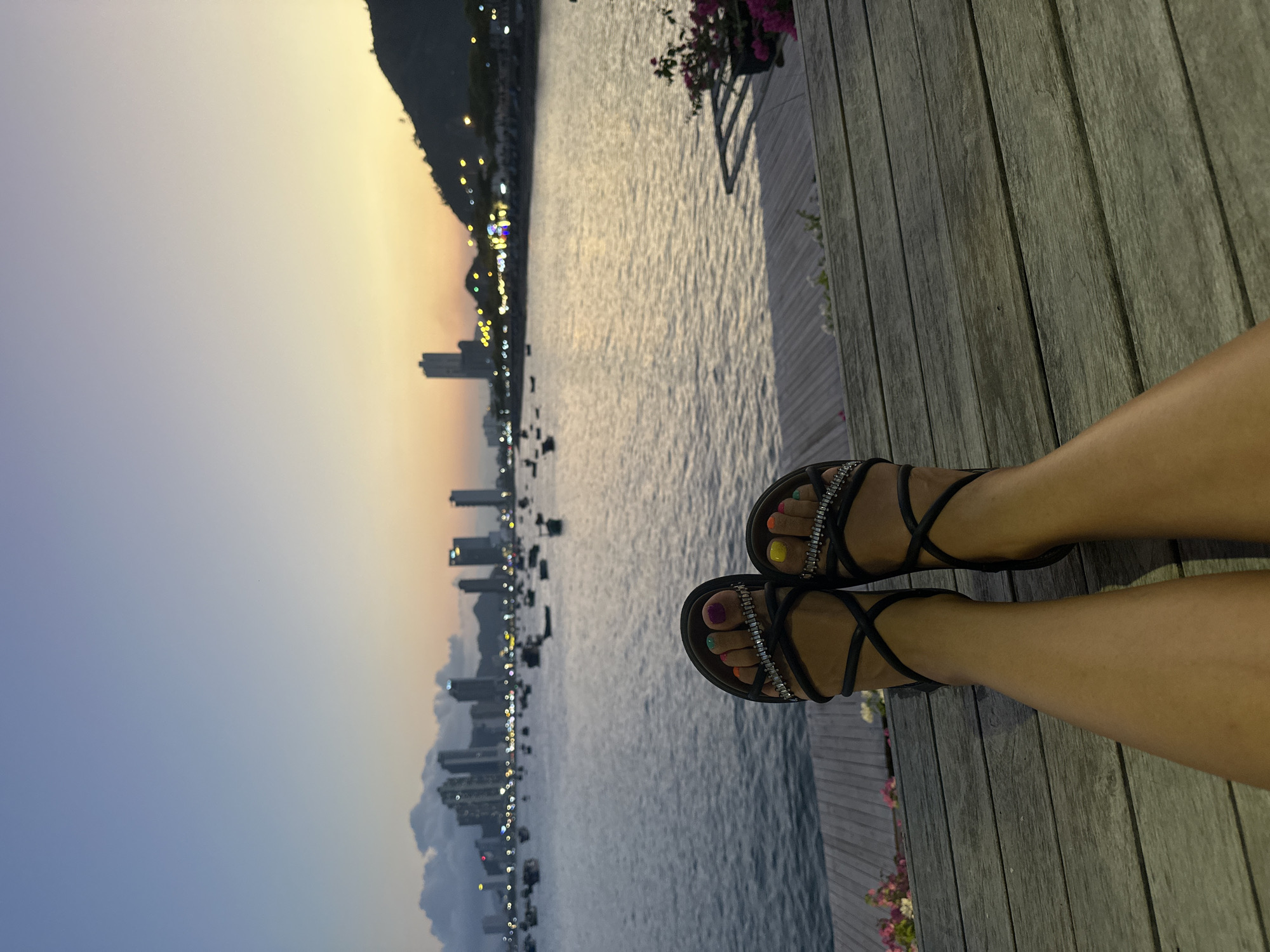 The image displays a coastal scene in Nha Trang, Vietnam, at dusk or early evening. In the immediate foreground, the lower legs and feet of a single individual are visible, resting on a greyish-brown wooden plank deck. The individual wears black strappy sandals adorned with rhinestone embellishments. Each toe is painted a distinct, bright color, including purple, orange, green, pink, yellow, and blue. To the far right on the deck, a potted plant with pink flowers is partially visible.
Beyond the deck, a body of dark water, likely Nha Trang Bay, stretches across the mid-ground, reflecting the twilight sky. Numerous small boats are scattered across the water. The background reveals the Nha Trang cityscape along the far shore, characterized by multiple high-rise buildings, some with illuminated windows or rooftops. A brightly lit road or shoreline runs along the base of the city and continues towards the right where a dark, vegetated hill or mountain rises. The sky transitions from a light blue overhead to a warm orange and yellow hue at the horizon, particularly on the right side.