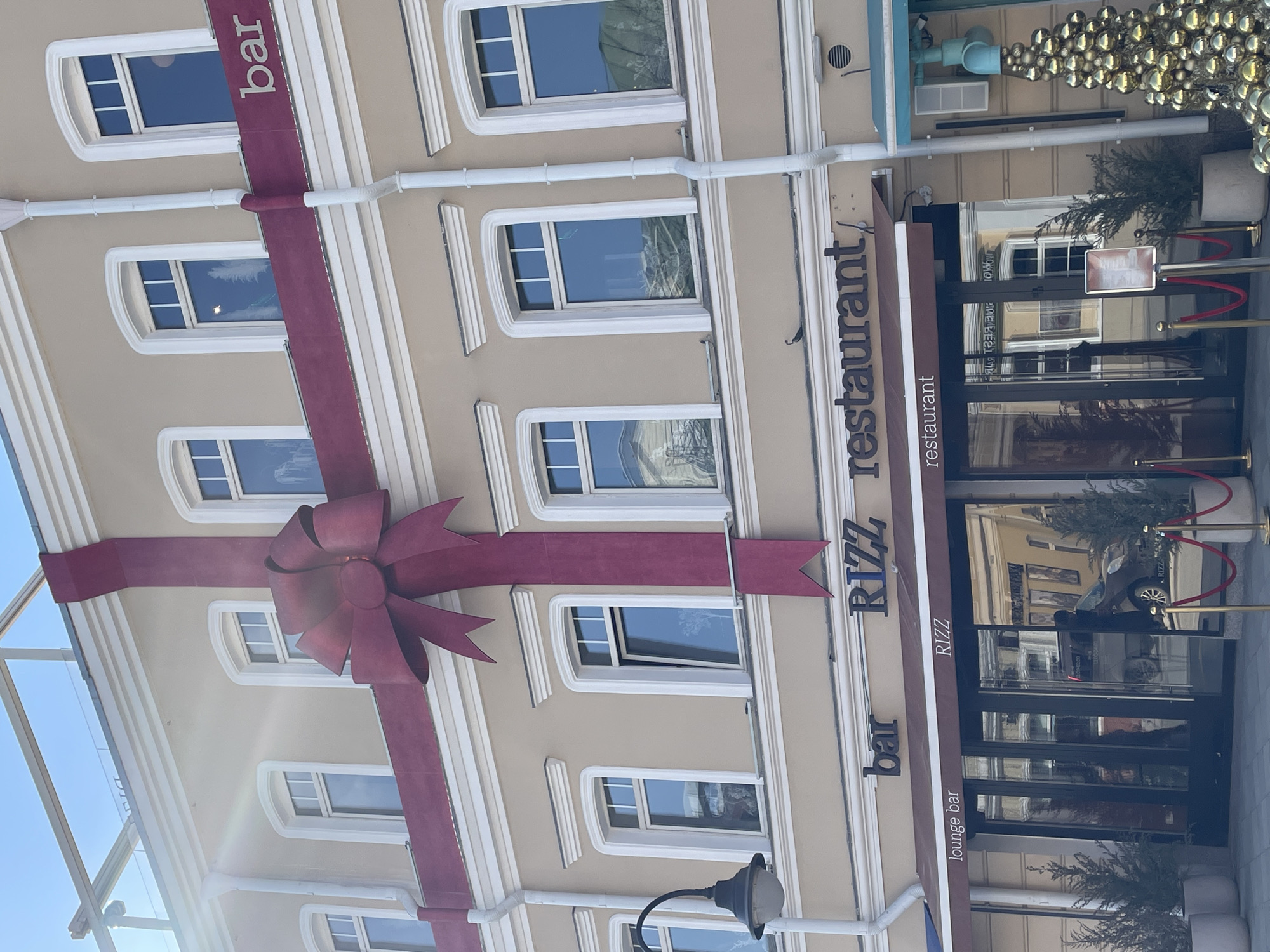 The image displays the facade of a three-story building, identified as "RIZZ restaurant" and "bar," situated on Tverskaya Street in Moscow, Russia. The building's light beige stucco exterior is decorated with a prominent, dark red ribbon and a large central bow, simulating a wrapped gift. The ribbon extends both vertically and horizontally across the facade. Multiple rectangular windows with white frames punctuate the upper floors, reflecting a clear blue sky and portions of the urban environment. A white downspout is present on the left side of the facade.
The ground floor features a dark brown awning labeled "bar RIZZ restaurant" in dark lettering, with additional signage for "lounge bar" and "RIZZ restaurant" in white lettering below. The glass entrance doors and windows reflect a dark vehicle and a human silhouette. The entrance area is flanked by potted evergreen trees and delineated by red velvet ropes and golden stanchions. A menu stand is positioned near the entrance. To the right of the entrance, a conical outdoor decoration composed of numerous gold spherical ornaments is visible. A dark street lamp is affixed to the building's left side. The scene is illuminated by bright sunlight, generating a lens flare in the upper left quadrant.