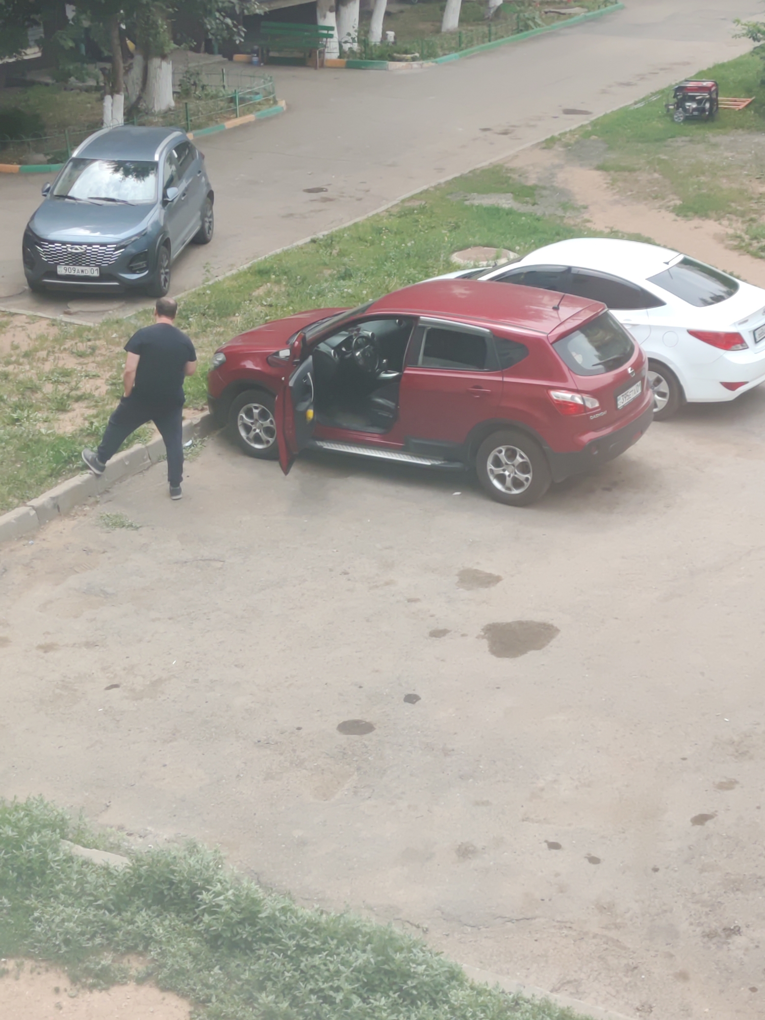 A man in a black t-shirt and pants is standing on a concrete curb next to a red SUV with its driver's side door open. The SUV is parked on asphalt. Behind the SUV, a white sedan is parked. In the background, a grey SUV is parked on a paved surface. There is also a portable generator on a grassy area to the right of the white sedan. The scene appears to be a residential area in Astana, Kazakhstan, with trees and some greenery visible in the background.