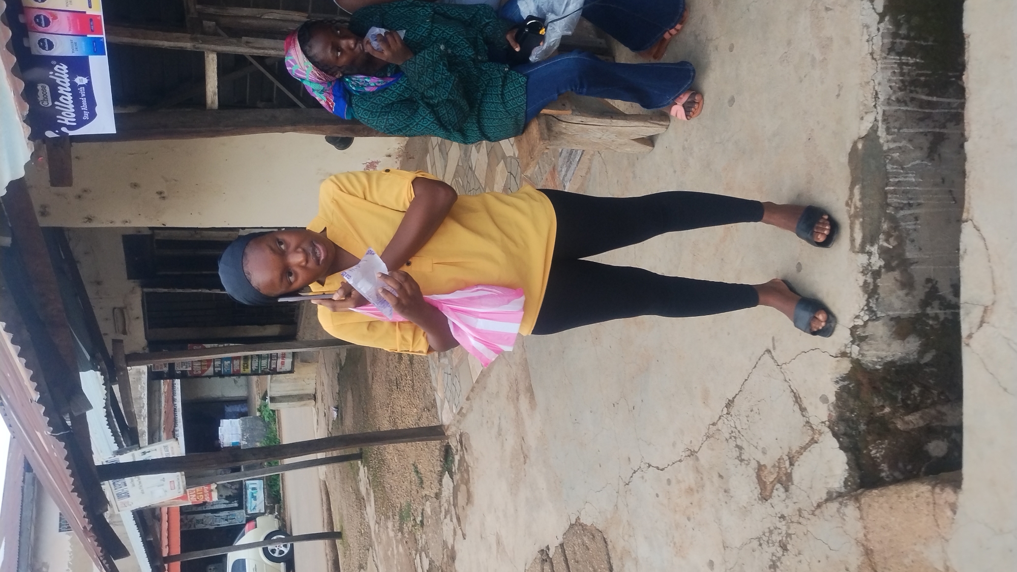This media file depicts an outdoor street scene in Jos, Nigeria, featuring two young women and elements of a local marketplace or roadside area.
In the foreground, a young woman stands, facing slightly towards the viewer. She is dressed in a bright yellow long-sleeved shirt, black leggings, and black slide sandals. A dark head covering completes her attire. She holds a mobile phone in her left hand and a white and pink striped plastic bag in her right, possibly looking at her phone or the camera.
Behind her, another young woman is seated on a low wooden bench. She wears a dark green patterned long-sleeved top, blue jeans, and pink slide sandals, with a colorful headscarf wrapped around her head. She holds a small white bottle or container.
The setting includes visible wooden support beams and corrugated metal roofing, suggesting a covered stall or entrance to a shop. A prominent sign is displayed on the left, featuring the brand name "Hollandia" with the text "Choose" above it and "Stay Ahead with" below. Other indistinct signs and partially visible buildings line the street in the background, along with part of a white vehicle. The ground is paved with cracked concrete and appears somewhat weathered. The scene is illuminated by daylight, indicating a clear morning or afternoon. The overall atmosphere is casual and reflects everyday life in an urban environment.
