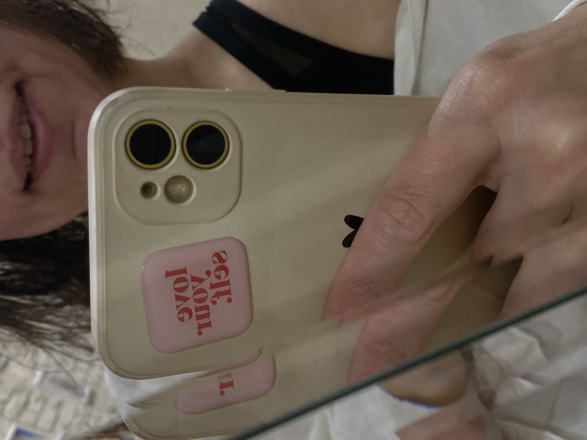 The image shows a person holding a beige smartphone with a dual-lens camera setup and a light pink sticker that says "Self Love" in reverse. The person has dark hair and a visible portion of their face, including a smile. They are wearing a black garment with mesh detailing. The background appears to be a reflection in a mirror or a glass surface. The environment seems to be a personal space, possibly a bedroom. The location of Yoshkar-Ola, Russia, is not verifiable from the image alone.