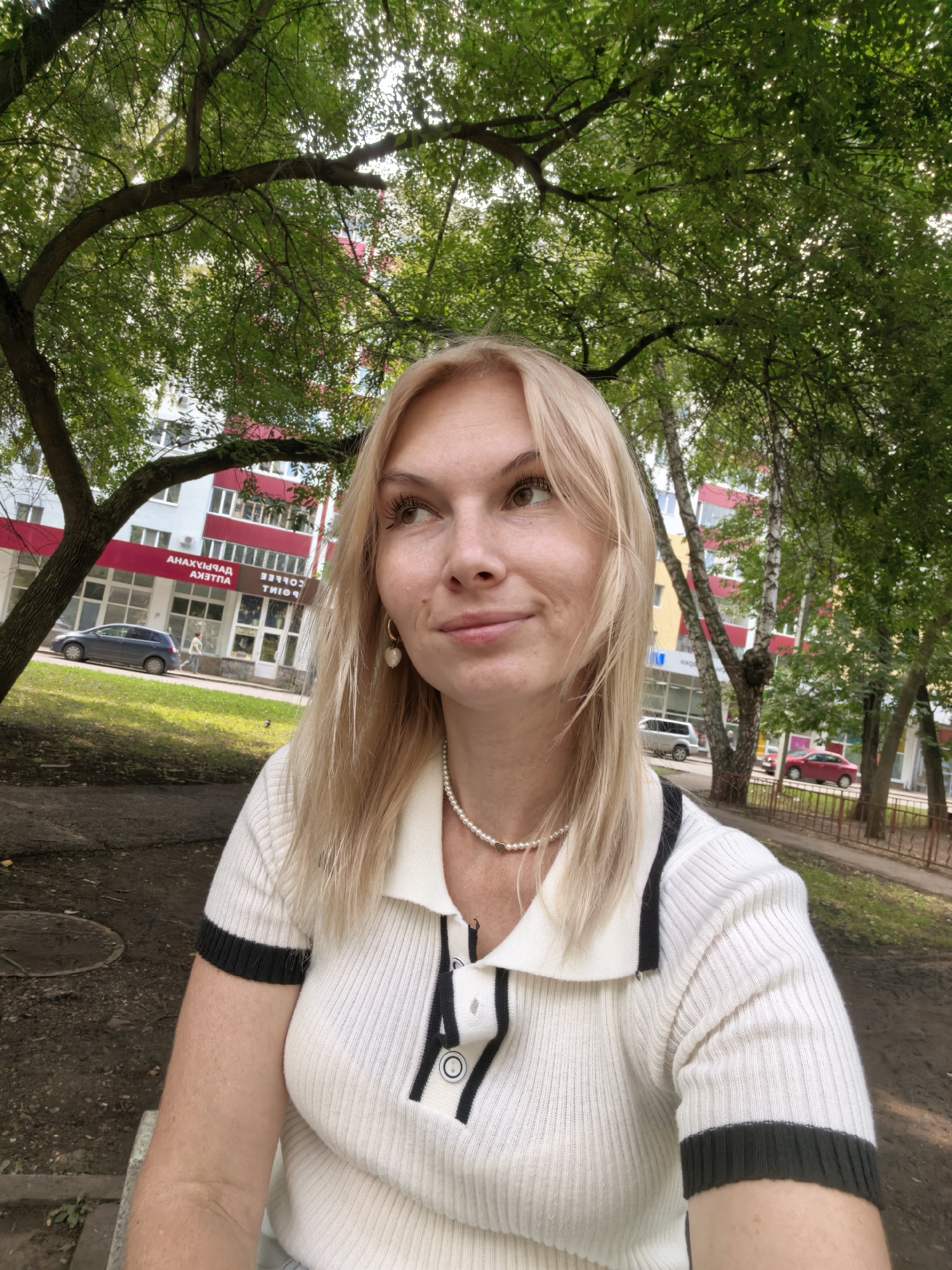 A young blonde woman with fair skin and light brown eyes is pictured in a selfie taken outdoors. She is wearing a white collared shirt with black trim on the collar and sleeves, and a pearl necklace. She has a light expression. Behind her, a building with white and red accents is visible, and trees with green leaves provide a natural backdrop. The scene appears to be in a residential area, possibly a courtyard or park. The woman's posture and the background suggest a casual outdoor setting.