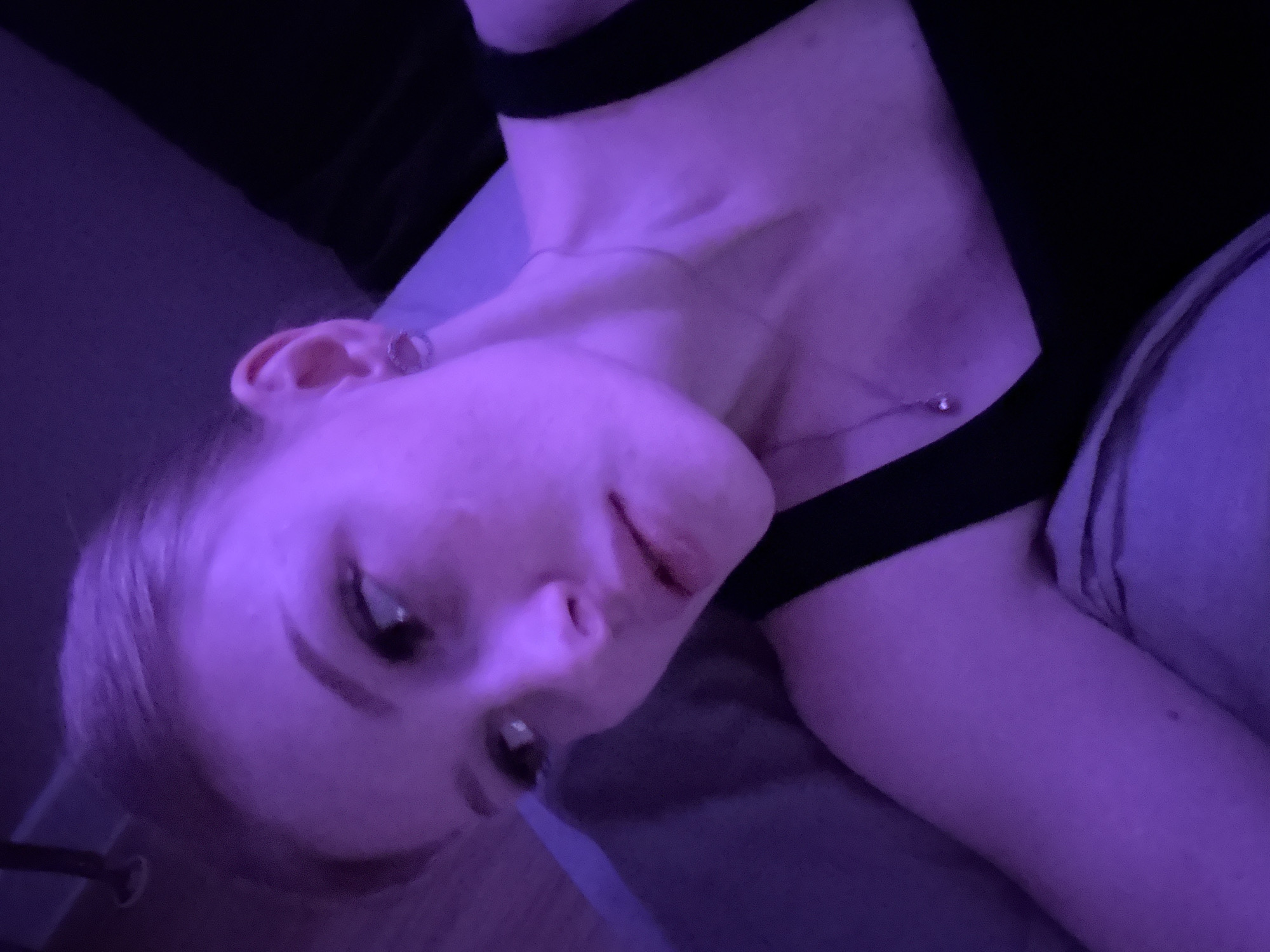 A young, fair-skinned woman is lying down indoors, possibly on a bed, viewed from a slightly high angle. She has light blonde hair and is wearing a black top with thick straps. A small silver necklace is visible around her neck, and she has an earring in her left ear. The room is illuminated with purple light. The background is dark and indistinct, with a portion of what appears to be a headboard or wall visible in the upper left. The subject’s gaze is directed downwards. The location in Perm, Russia, is not discernible from the image.