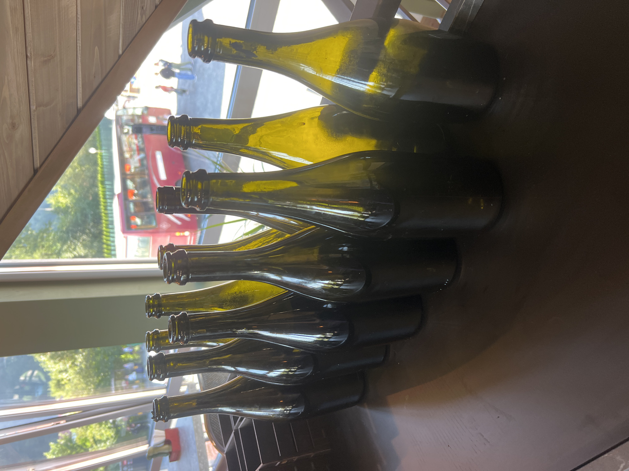 A group of ten empty, dark green glass bottles stand in a line on a dark wooden surface. The bottles are all of similar shape and size, with narrow necks and wide bases. They are arranged in two rows, with the front row of bottles slightly overlapping the bottles in the back row. The surface they are on appears to be a bar or counter.
In the background, large windows overlook an outdoor scene. Sunlight streams through the windows, casting reflections on the bottles. Outside, trees with green foliage are visible, along with a red bus and several people walking. The setting appears to be a cafe or restaurant with large windows.
The city of Yaremche, Ukraine, is known for its natural beauty and is a popular tourist destination, with many cafes and restaurants offering views of the surrounding mountains and forests. The scene depicted in the image is consistent with such a location.