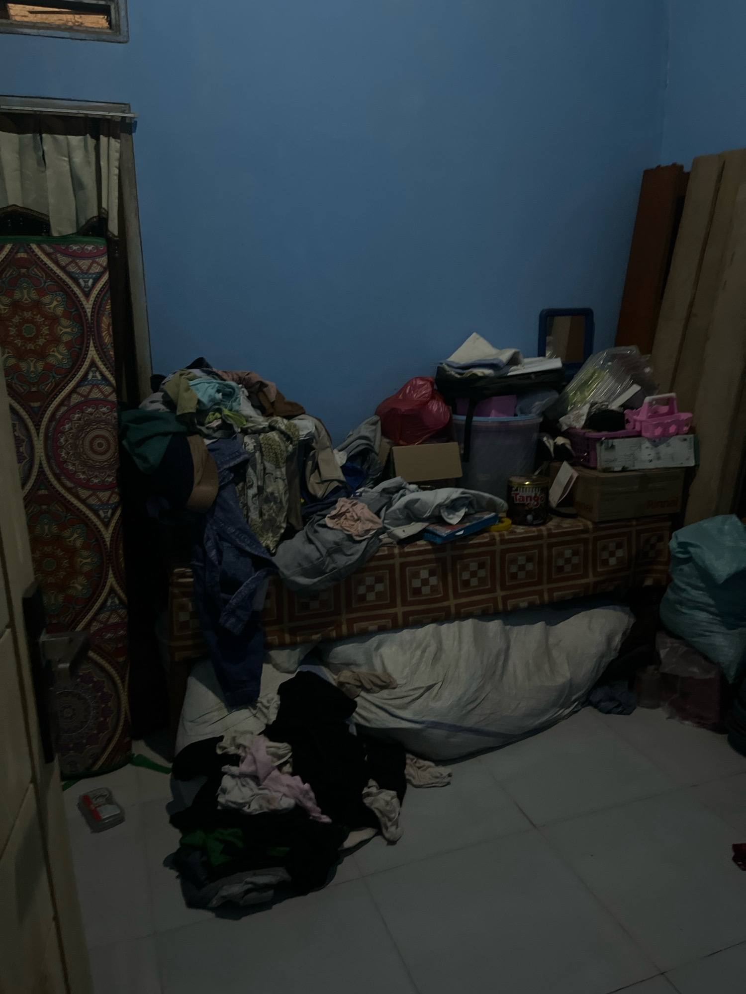 The image depicts a cluttered room, possibly a bedroom, in Kebumen, Indonesia. The walls are painted a light blue. On the left side of the room, a large, ornate tapestry hangs vertically. In the center, a bed or couch is covered with a patterned cloth, and on top of it, a large pile of assorted clothing and items is haphazardly arranged. These items include what appear to be shirts, pants, and possibly some bags. There are also boxes, plastic containers, and a red bag visible within this pile. To the right of the bed, several wooden planks are leaning against the wall. In the foreground, more clothing is scattered on the floor, including a pile of dark-colored garments and lighter-colored items. A large, light-colored sack is also present on the floor. The overall impression is one of disarray and a lack of organization within the living space. The lighting suggests it is either dusk or an interior with dim illumination.