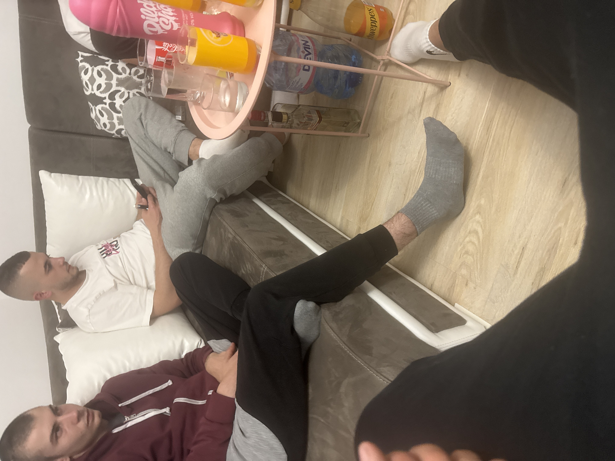 Three men are relaxing indoors. Two men are reclining on a couch, one wearing a white t-shirt and grey sweatpants, the other wearing a burgundy hoodie and grey sweatpants. The third man, partially visible, is wearing black pants. A side table with various beverages sits in front of the couch. The location is Graf Ignatievo, Bulgaria.