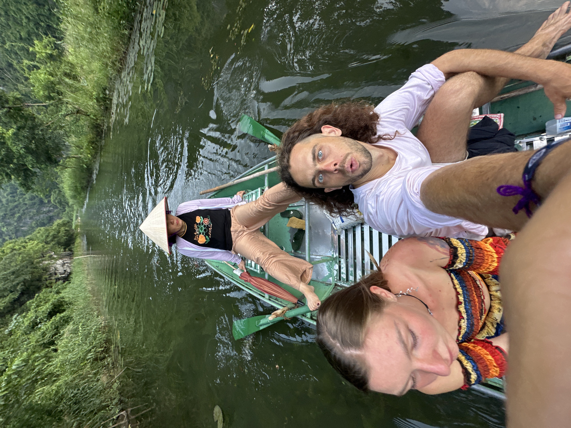 In Nam Hoa Lư Ward, Vietnam, three individuals are seated in a narrow boat traversing a calm, green body of water. The person in the rear wears a conical hat and a light purple jacket over a black shirt. In the middle section of the boat, a man with curly brown hair and blue eyes looks towards the camera with his mouth open. A woman with brown hair, wearing a colorful off-the-shoulder top, is positioned in the foreground, looking downwards. The surrounding environment is lush and green, with dense foliage lining the water's edge and tall, rocky cliffs in the background. The water is calm, reflecting the greenery and the sky.
